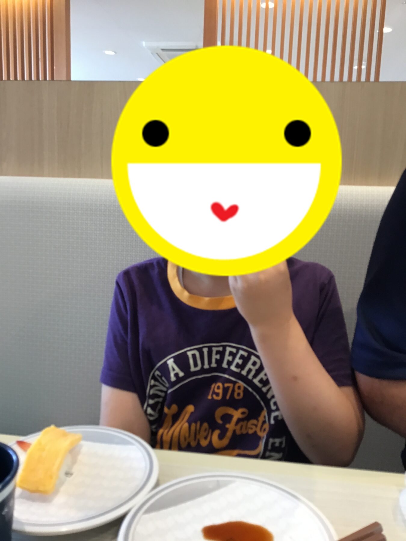 Child in a purple shirt sits at a restaurant table; a large yellow smiling emoji covers their face.