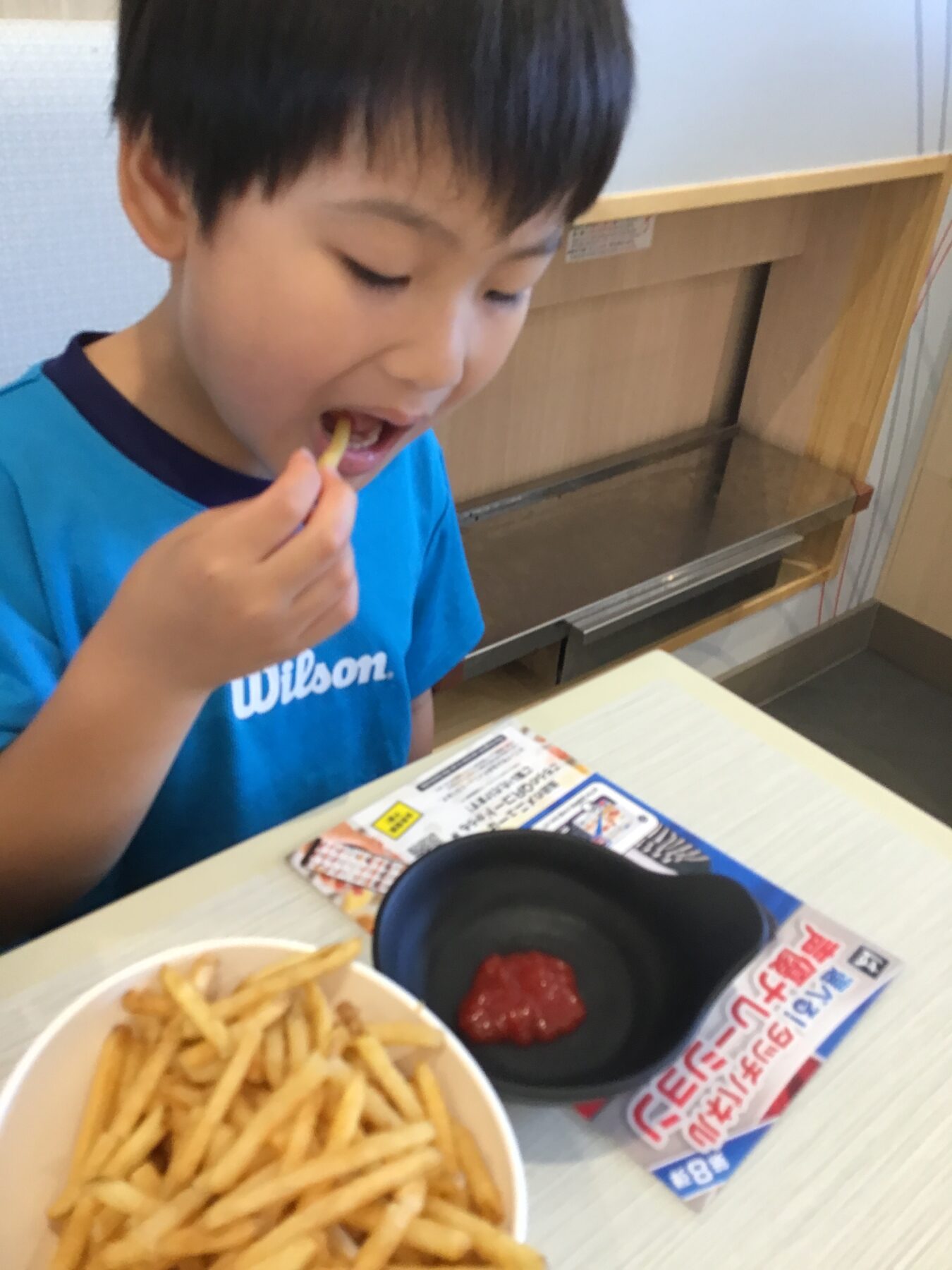 Child wearing a blue shirt biting into French fry at a table with fries and ketchup nearby in a casual dining setting