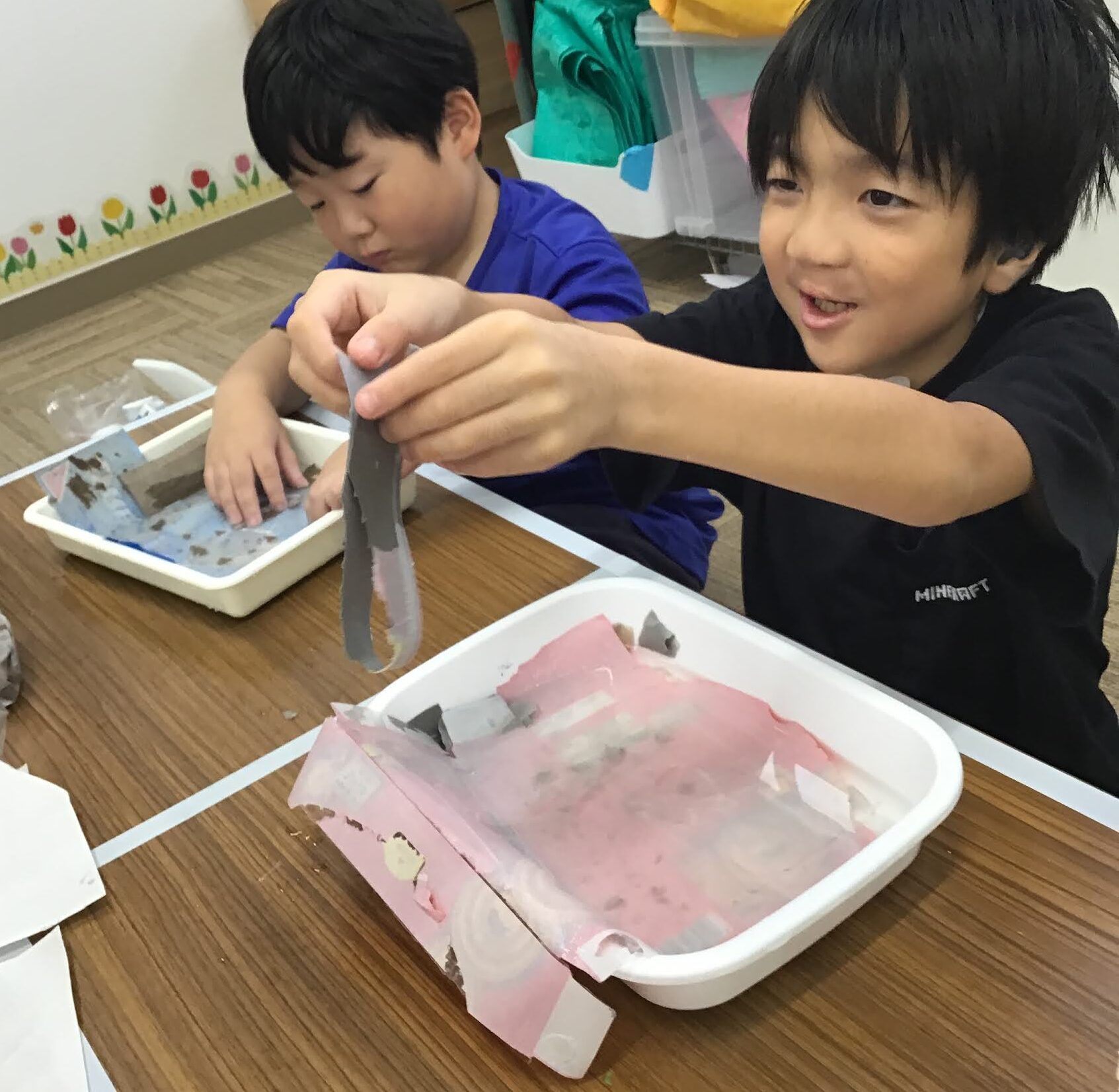 Two children at a table threading pink and gray paper strips for a craft project in a classroom setting.