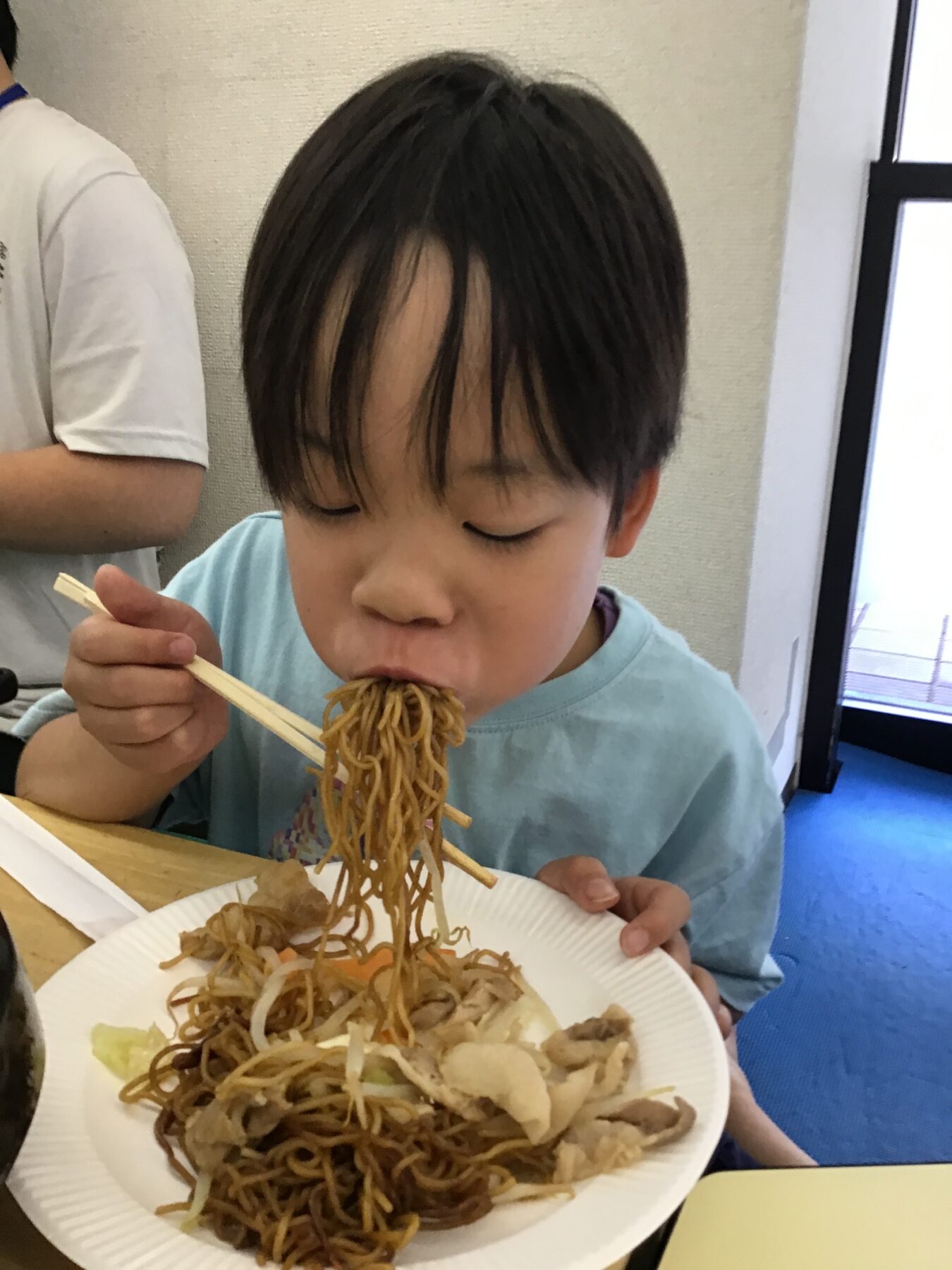 Young child wearing a light blue shirt eating noodles with chopsticks from a paper plate at a table.
