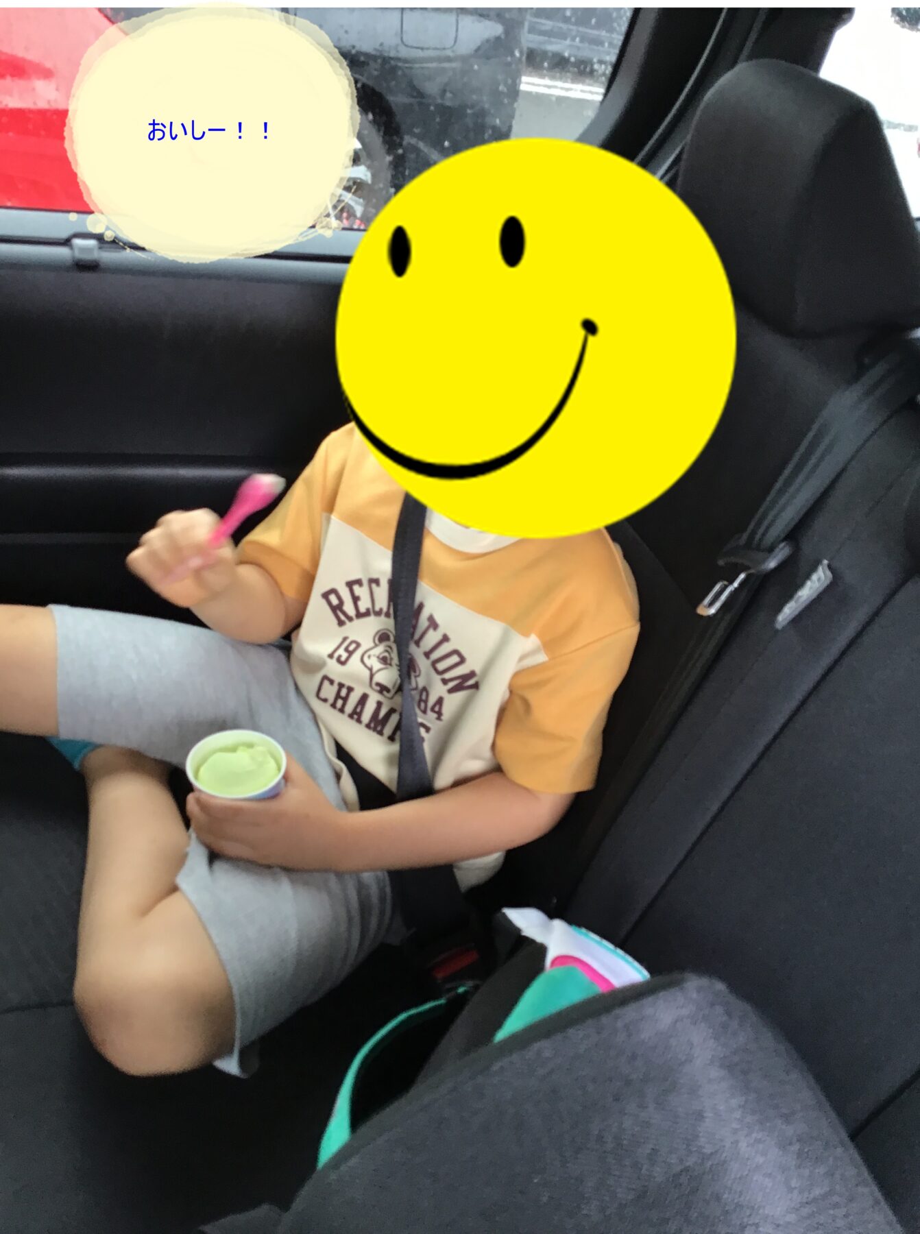 Child in back seat wearing an orange shirt, holding a cup; a large yellow smiley overlay covers their face with a blue speech bubble reading 'おいしー!!'.