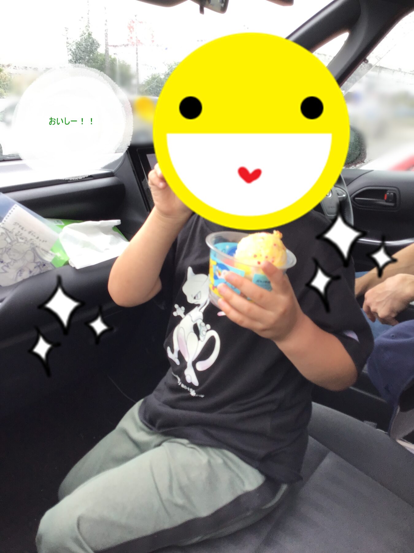Child in a car seat holding an icy cup of colorful ice cream, face covered by a large yellow smiley sticker with sparkles nearby.
