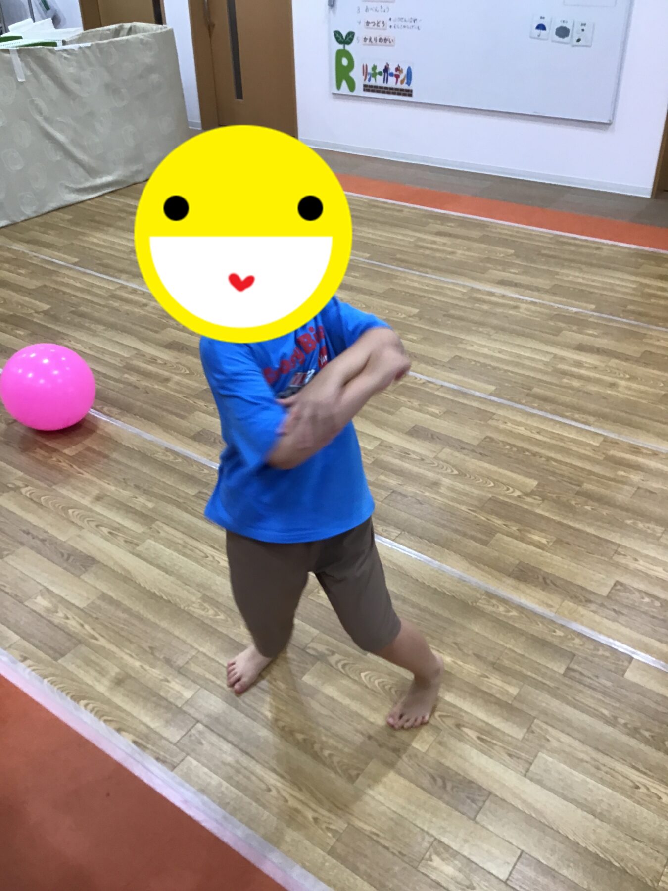 Child in a blue shirt and brown shorts, barefoot on a wooden gym floor, with a pink ball nearby; face covered by a large yellow smiley emoji.