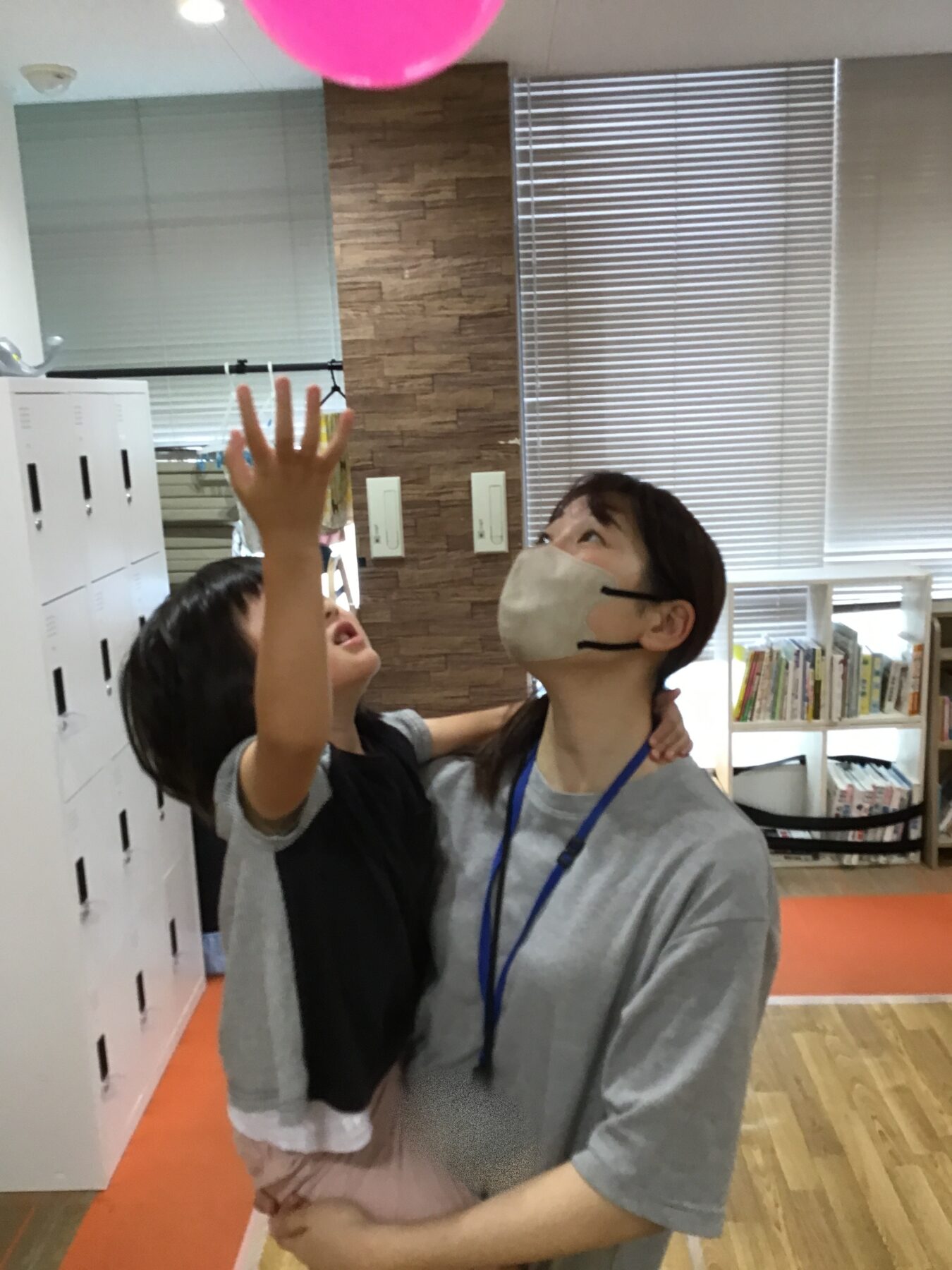 Two children indoors reach upward toward a pink balloon, one wearing a beige mask. A row of lockers and blinds are in the background.