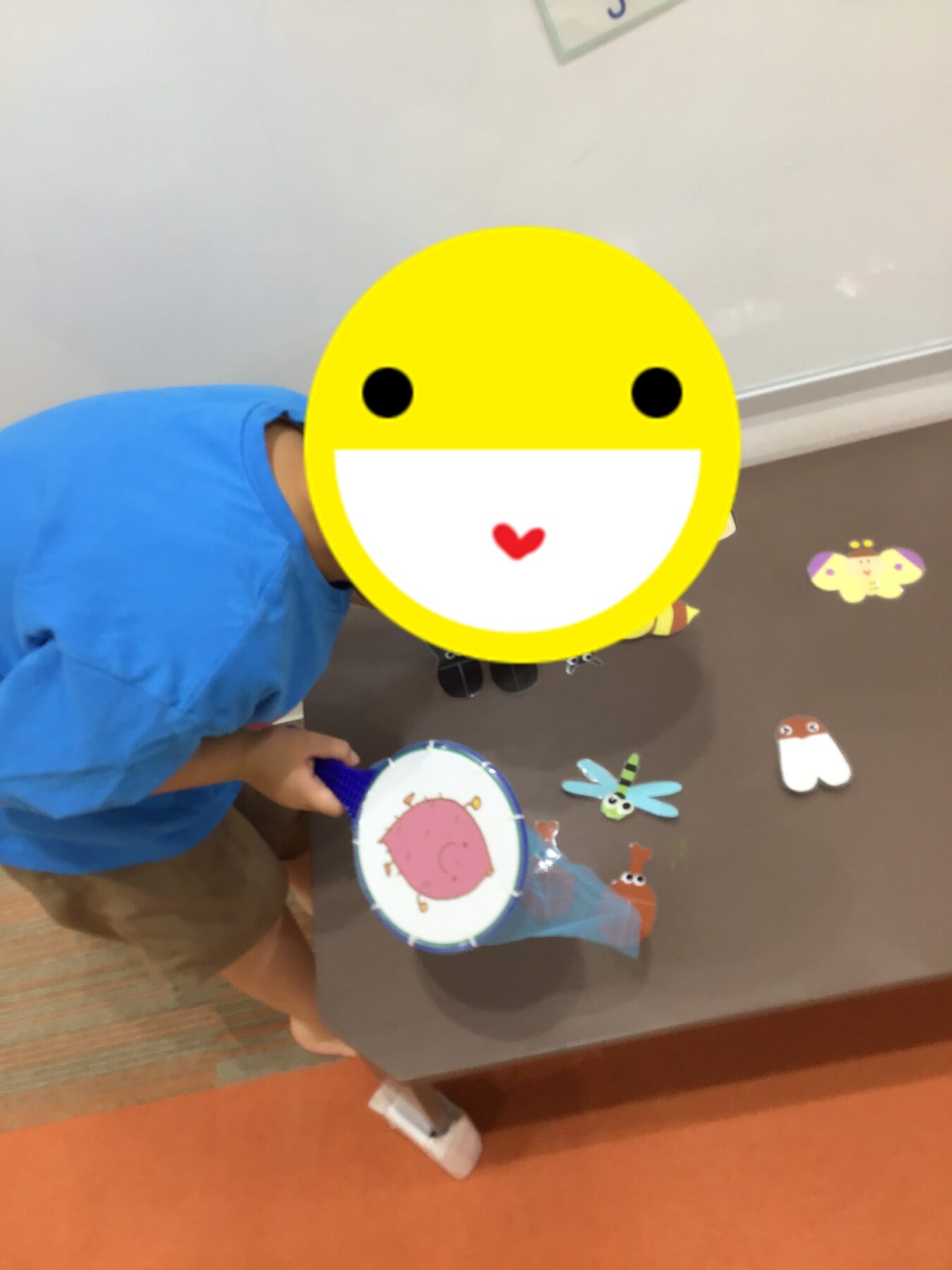 Child in a blue shirt playing with a small drum or musical toy on a table, with a large yellow smiley face sticker covering their face.
