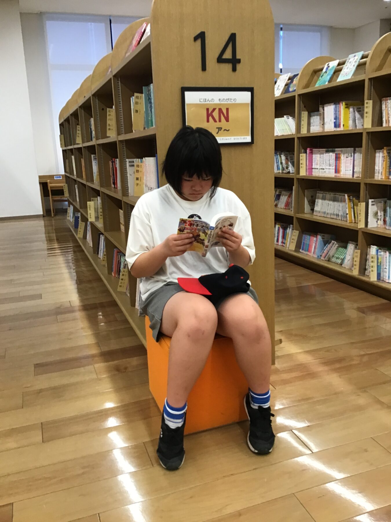 Child with black hair in a white shirt sits on an orange block, reading a small book in a library aisle with tall wooden shelves nearby.