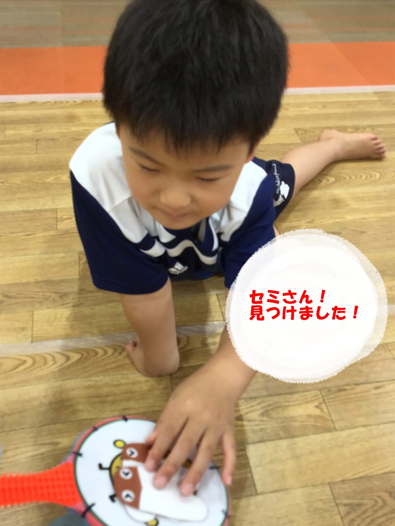 Boy in a blue and white shirt sits on a wooden floor, looking down at a round cartoon plate with a bear face, a white hat nearby and red Japanese text overlay reading 'セミさん!見つけました!'