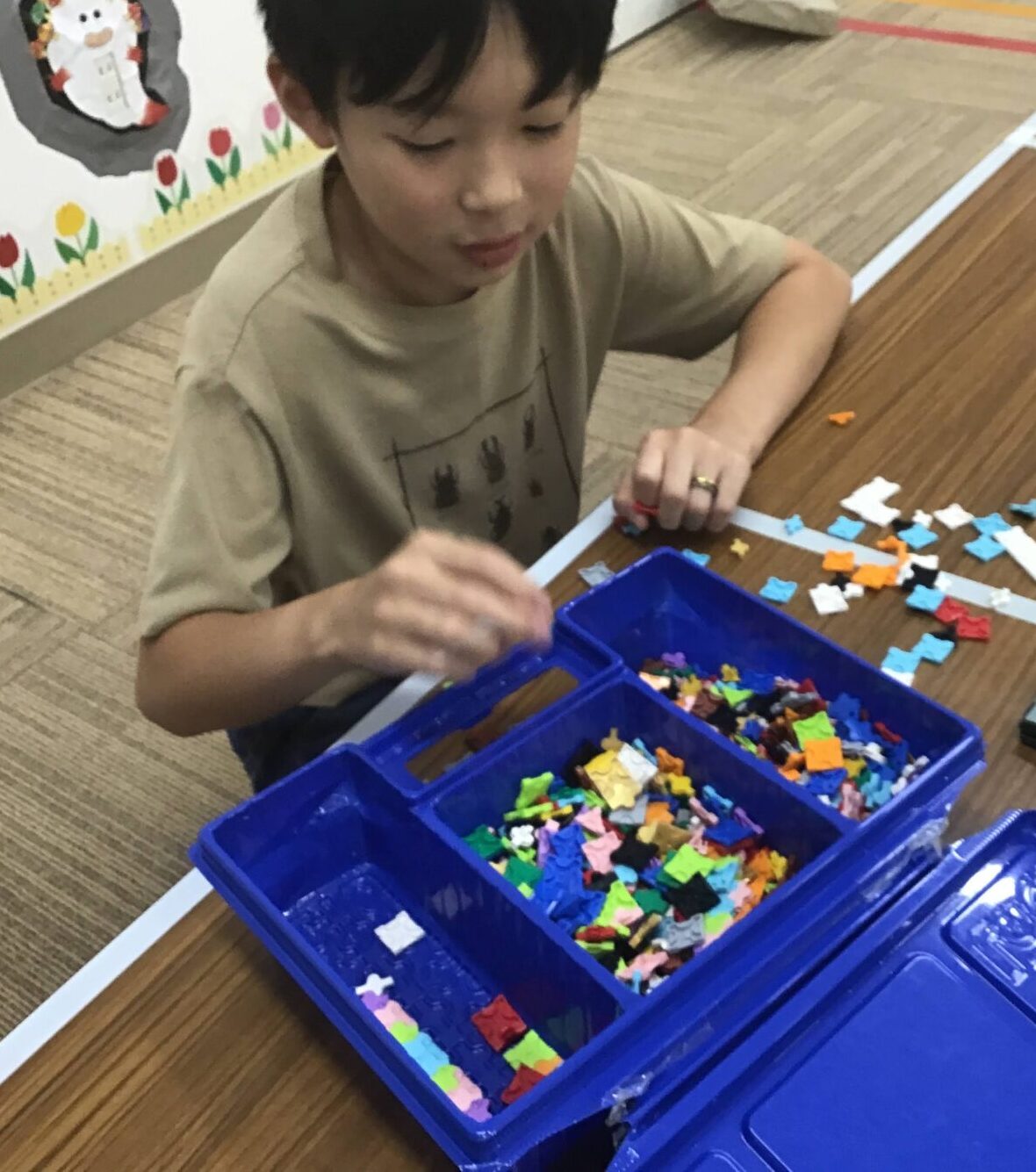 Boy assembling a mosaic with tiny colorful tiles in blue trays at a table; a €20 note sits nearby on the table.