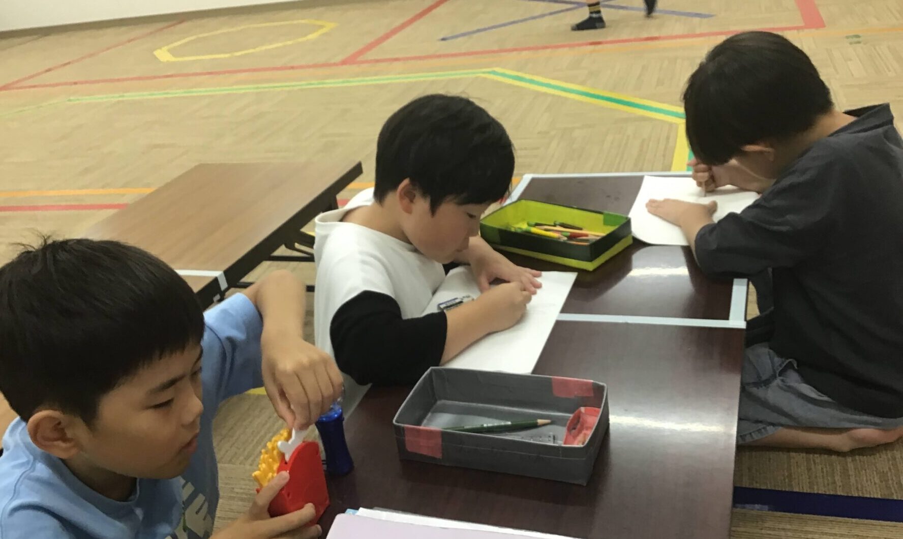 Children sit at a long table drawing and writing with pencils and paper in a gym-like room; colorful floor markings visible behind them.