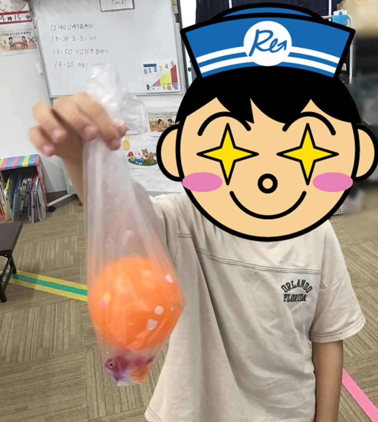 Child in a classroom holds up a clear plastic bag filled with an orange drink, with a playful sticker covering their face in the foreground.