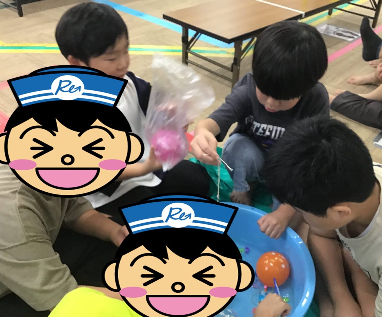 Children sit on the floor around a blue tray, threading colorful beads during a hands-on activity in a gym-like room.
