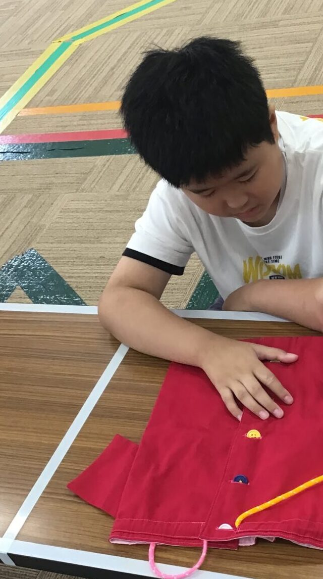 Boy sits at a classroom table sewing a red fabric with yellow trim and small colorful buttons visible on the cloth.
