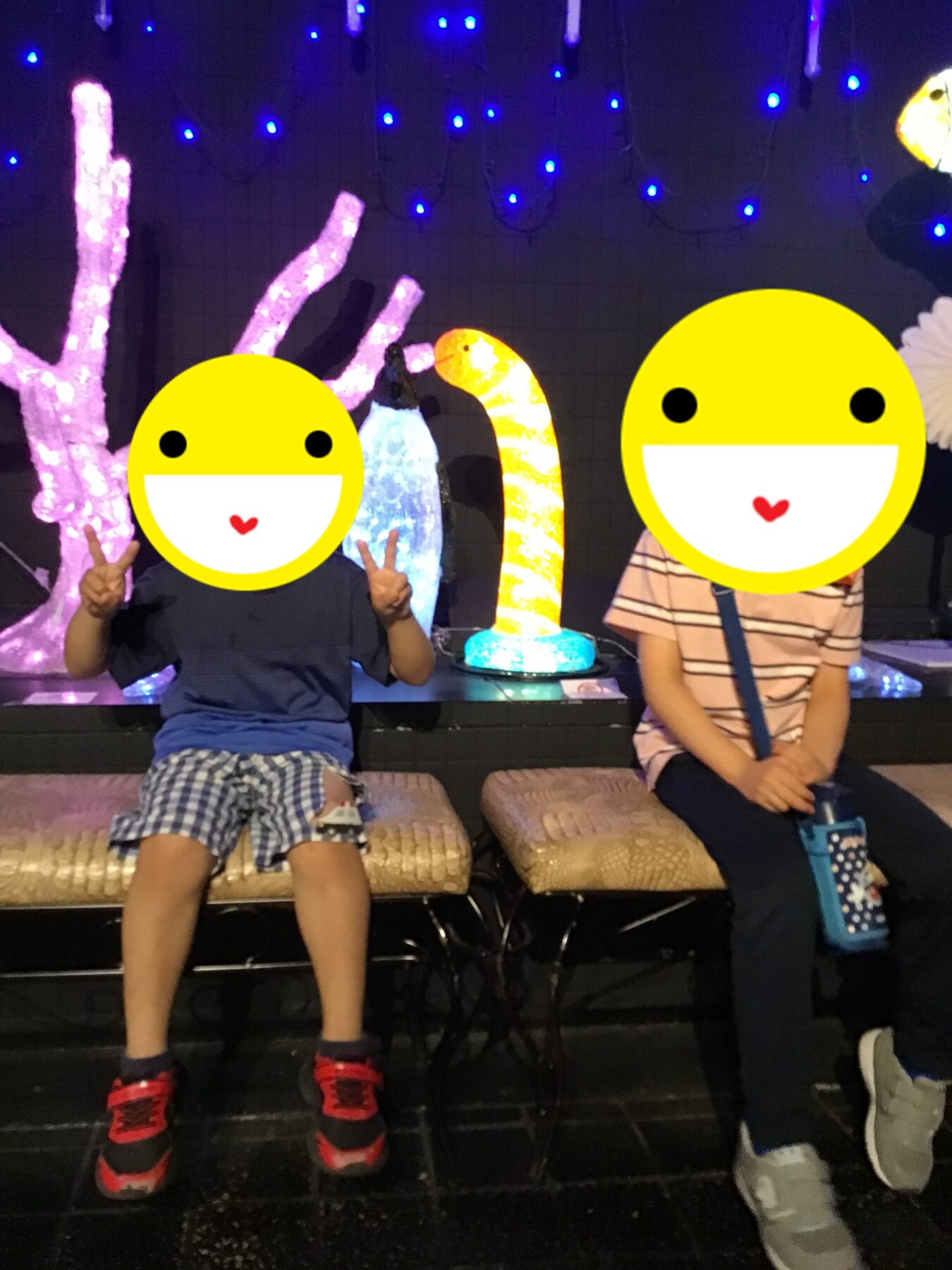 Two children sit on a bench with yellow smiley-face emoji overlays masking their faces in a lit, decorative exhibit backdrop.