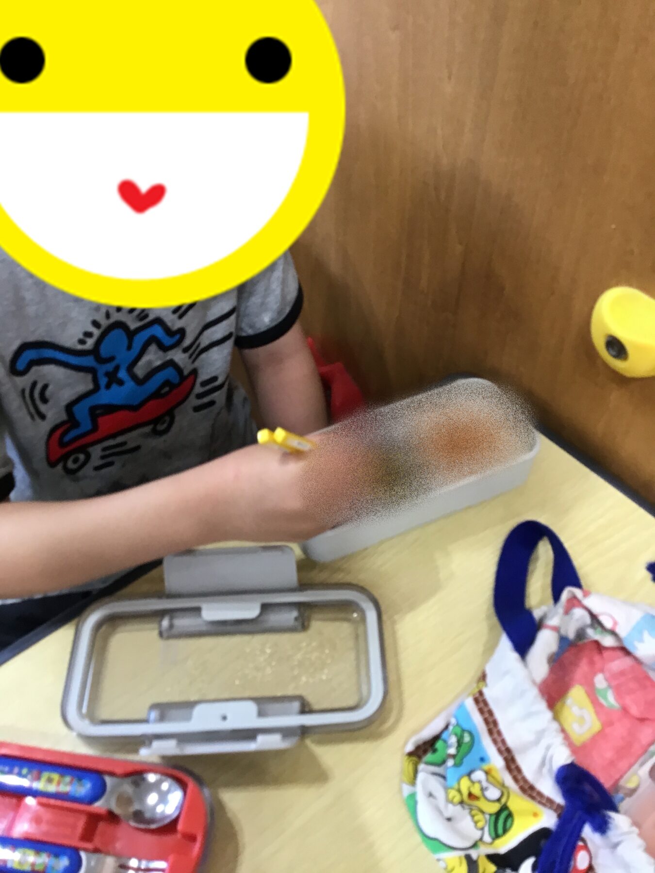Child with a large yellow emoji face sticker over their head, sitting at a table and drawing with a yellow pencil among craft supplies.