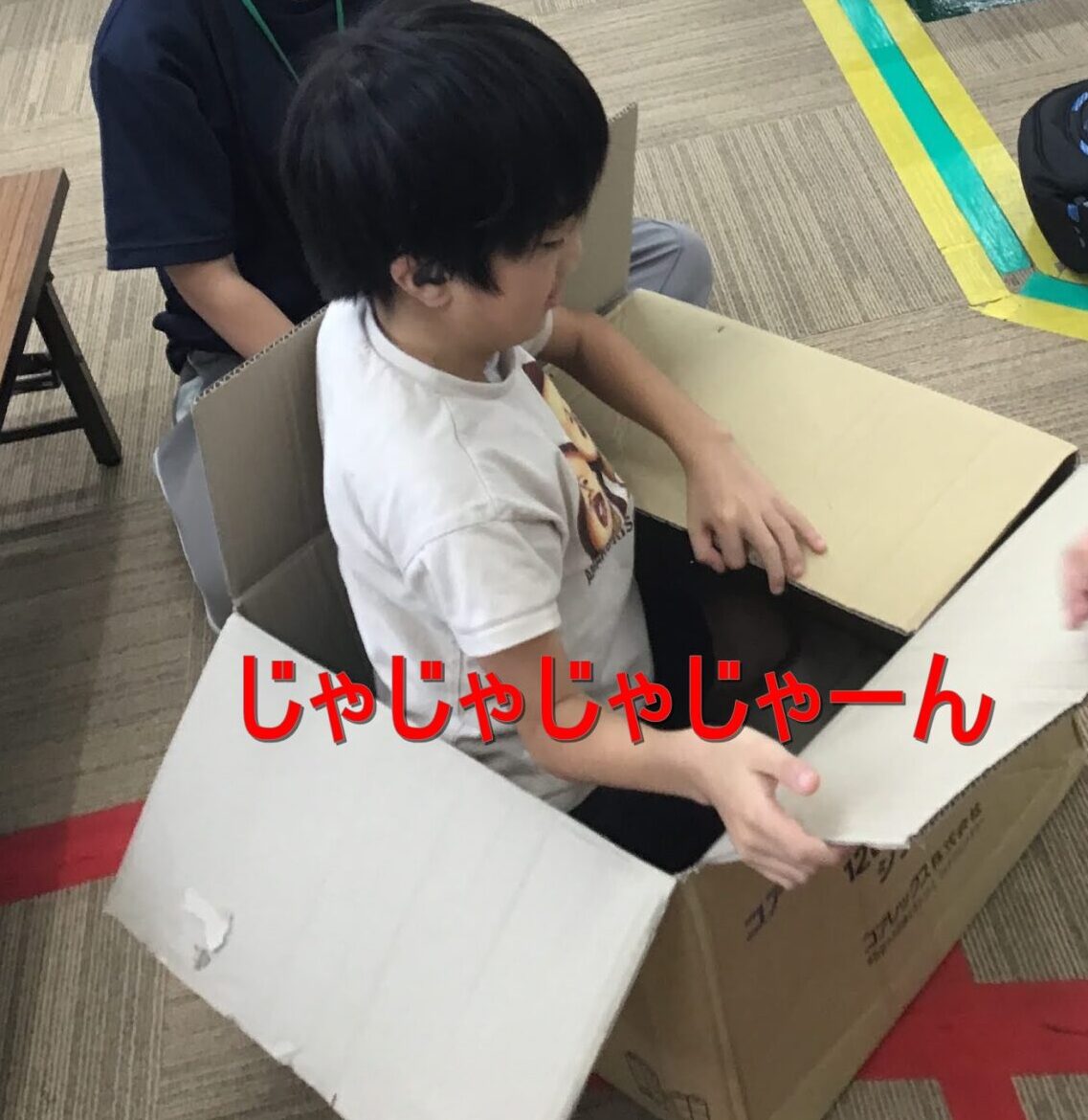 Two children in a classroom gym, one wearing a mask and glasses, sitting inside a large cardboard box as a friend stands nearby; colorful taped lines on the floor indicate a play area. The Japanese on-screen text “じゃじゃじゃじゃーん” appears in red at the bottom.