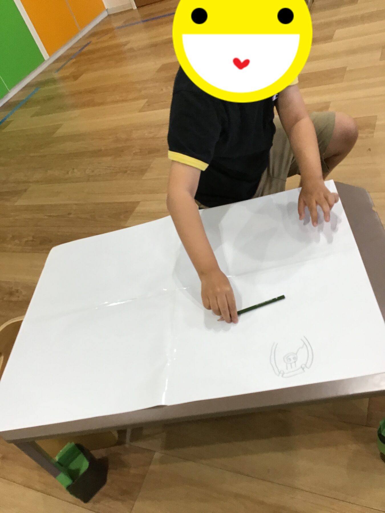 Child sitting at a white table, drawing with a green pencil; a large yellow emoji covers the face for privacy/fun.