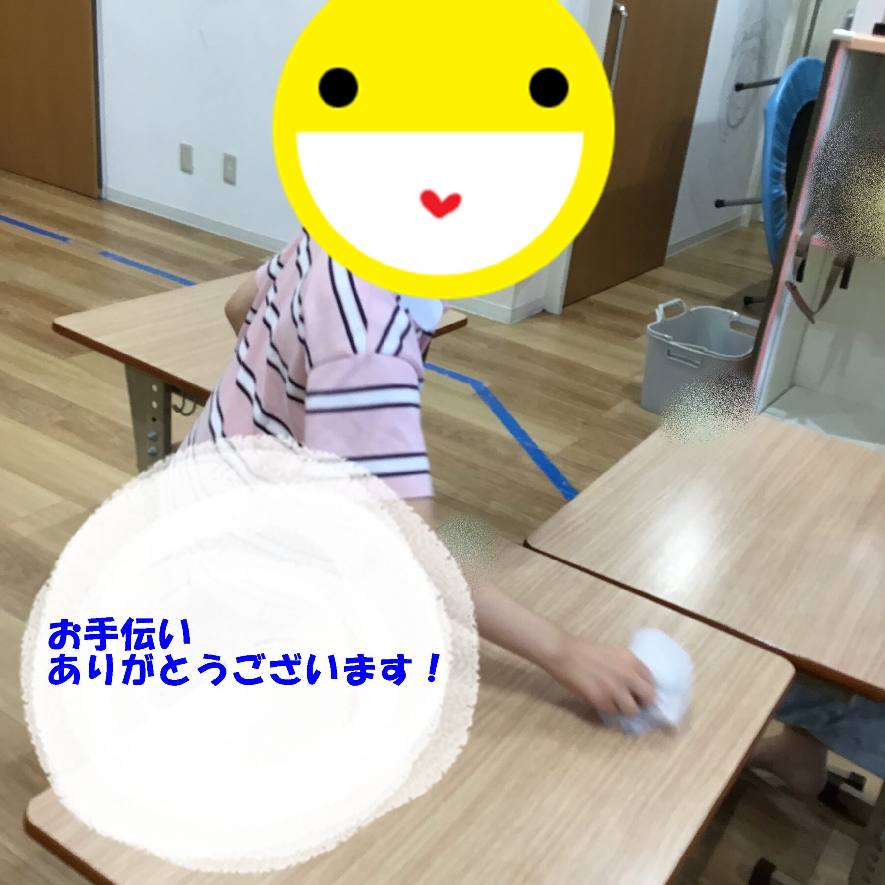 Child sits at a wooden table with a large yellow smiley face covering their own, blue Japanese text on a plate in the foreground reads 'お手伝いありがとうございます！'