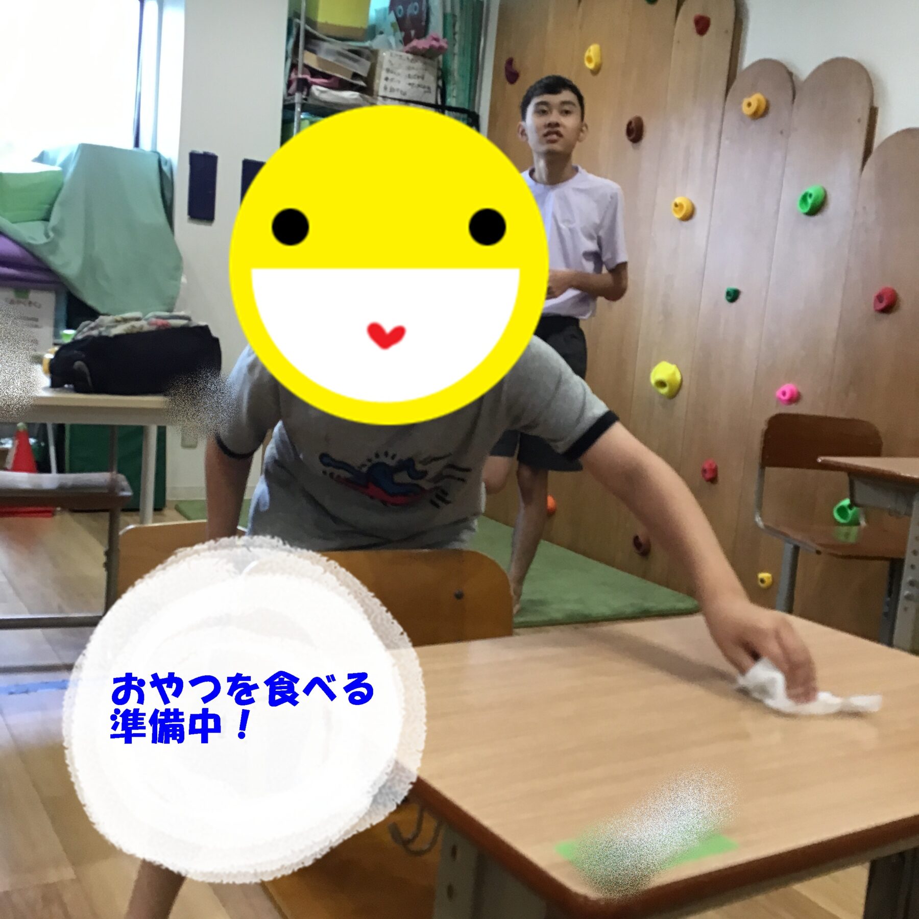 Child leaning on a table with a large yellow emoji covering their face; a climbing wall and other kids in the background in a classroom setting.