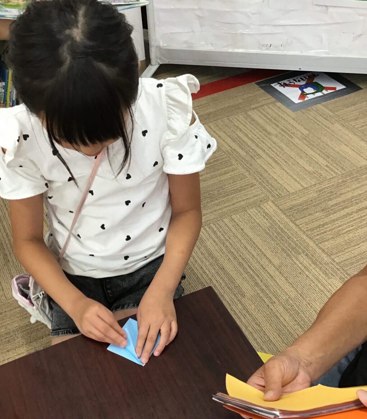 Young girl folding a blue origami square at a table in a classroom, with colorful papers nearby.