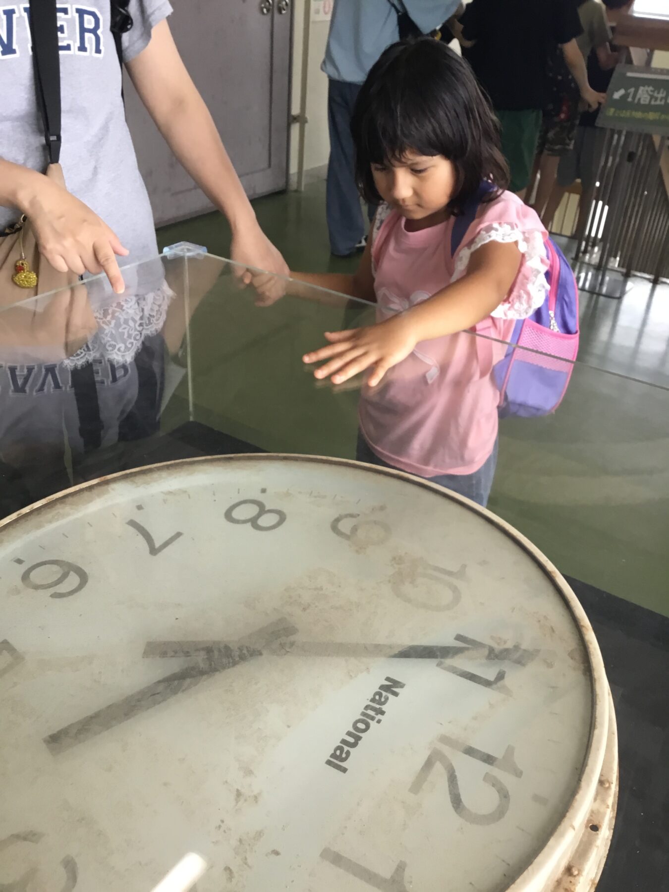 Young girl with a pink shirt and backpack touching a clear panel of an interactive exhibit, with a large circular dial in the foreground