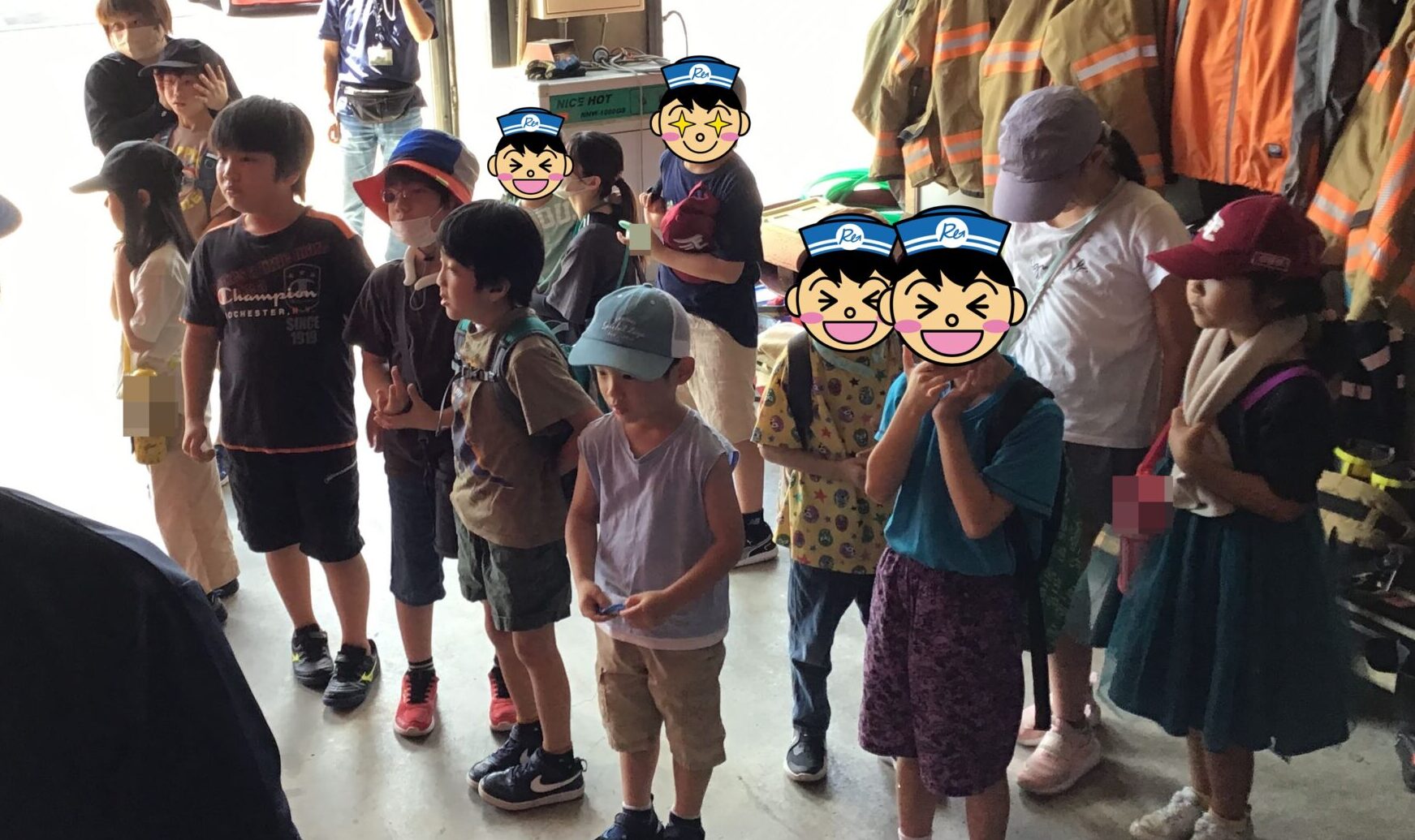 Group of children in a fire station bay listening to a guide, with turnout gear and helmets hanging on the wall behind them.