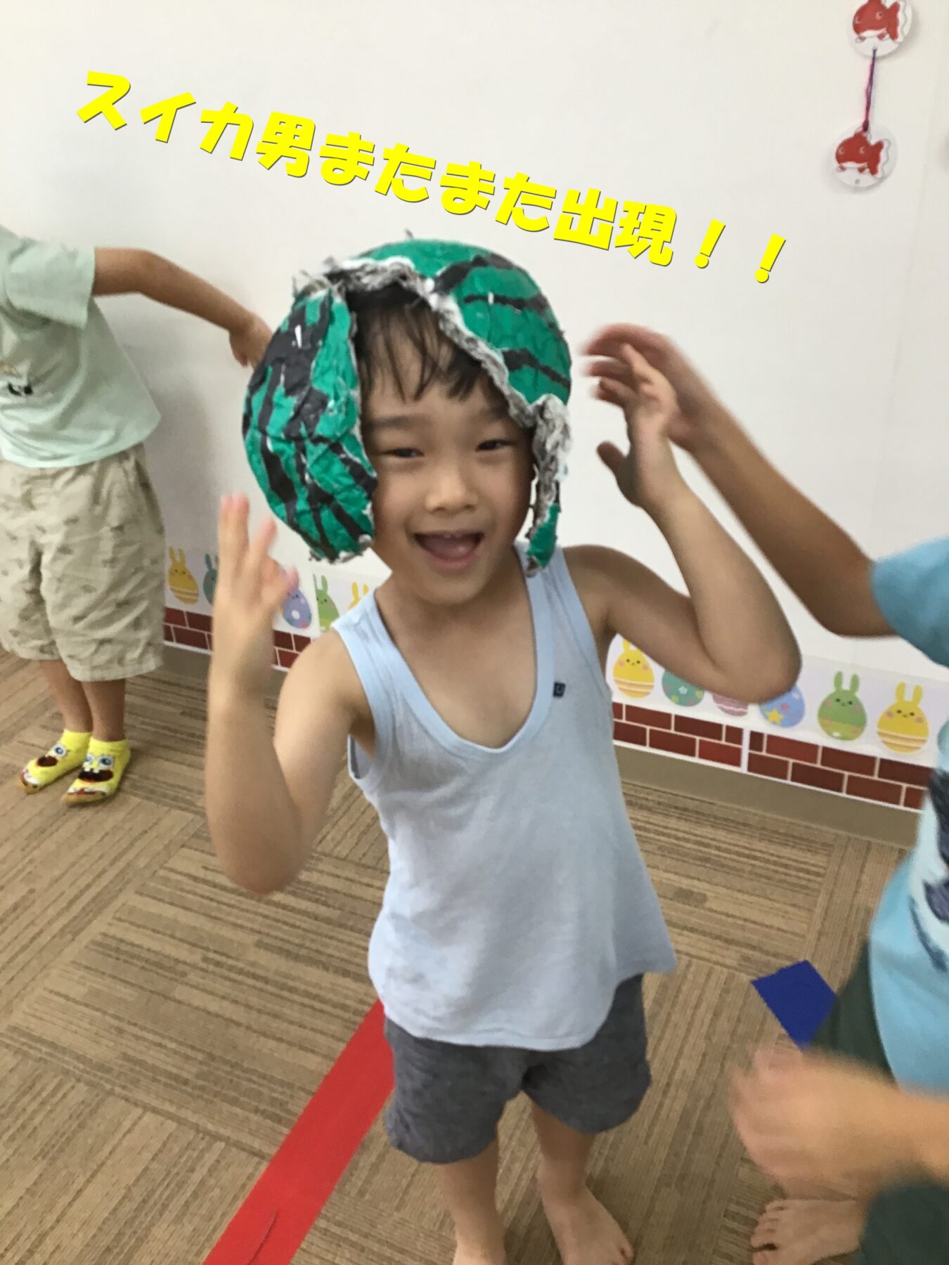 Young boy smiling while wearing a handmade green helmet, in a classroom with other kids nearby and playful decorations on the wall.