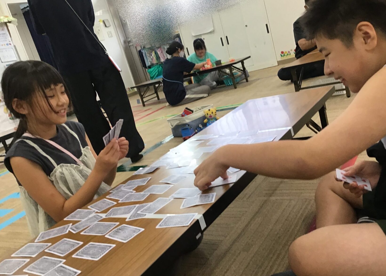 Two children sit across from each other at a low table, playing a card game with many cards spread out; the girl on the left smiles while holding her cards, and the boy on the right leans in with his cards in hand in a bright indoor room.