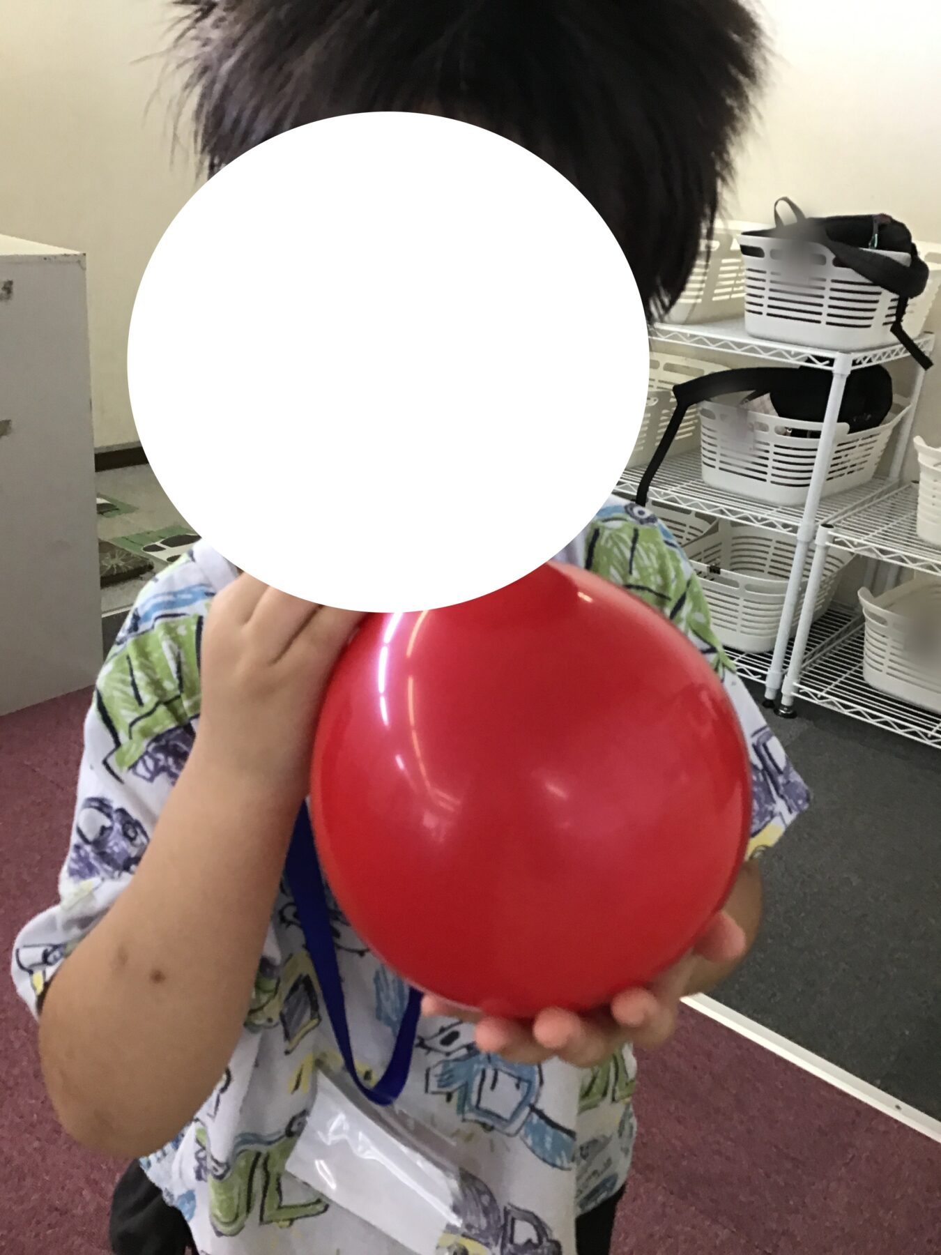 Child wearing a tropical shirt holds a bright red balloon in front of their face, with shelves and white baskets visible in the background.