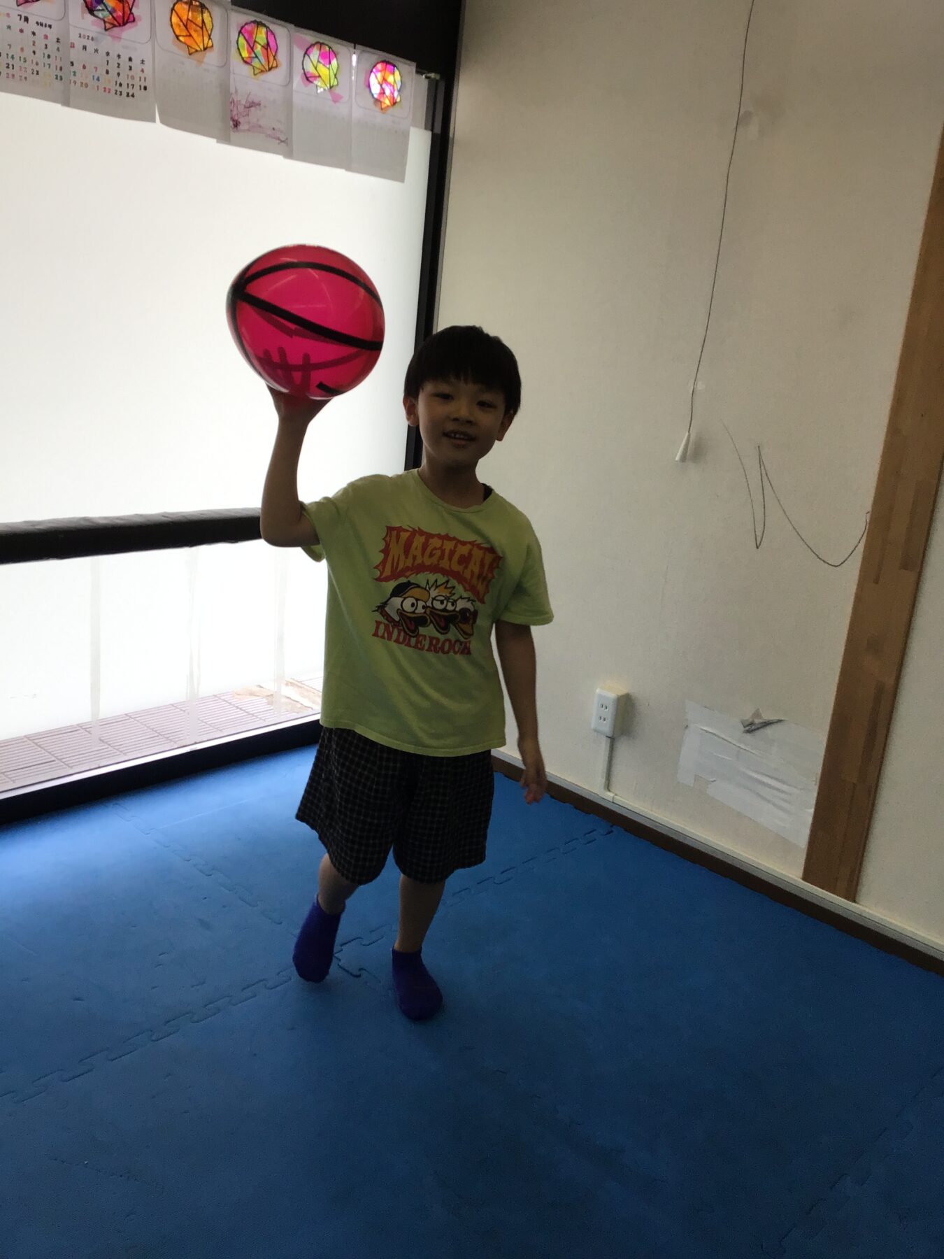 Young boy in a light green shirt and plaid shorts holds a bright pink basketball aloft in a room with blue padded flooring.