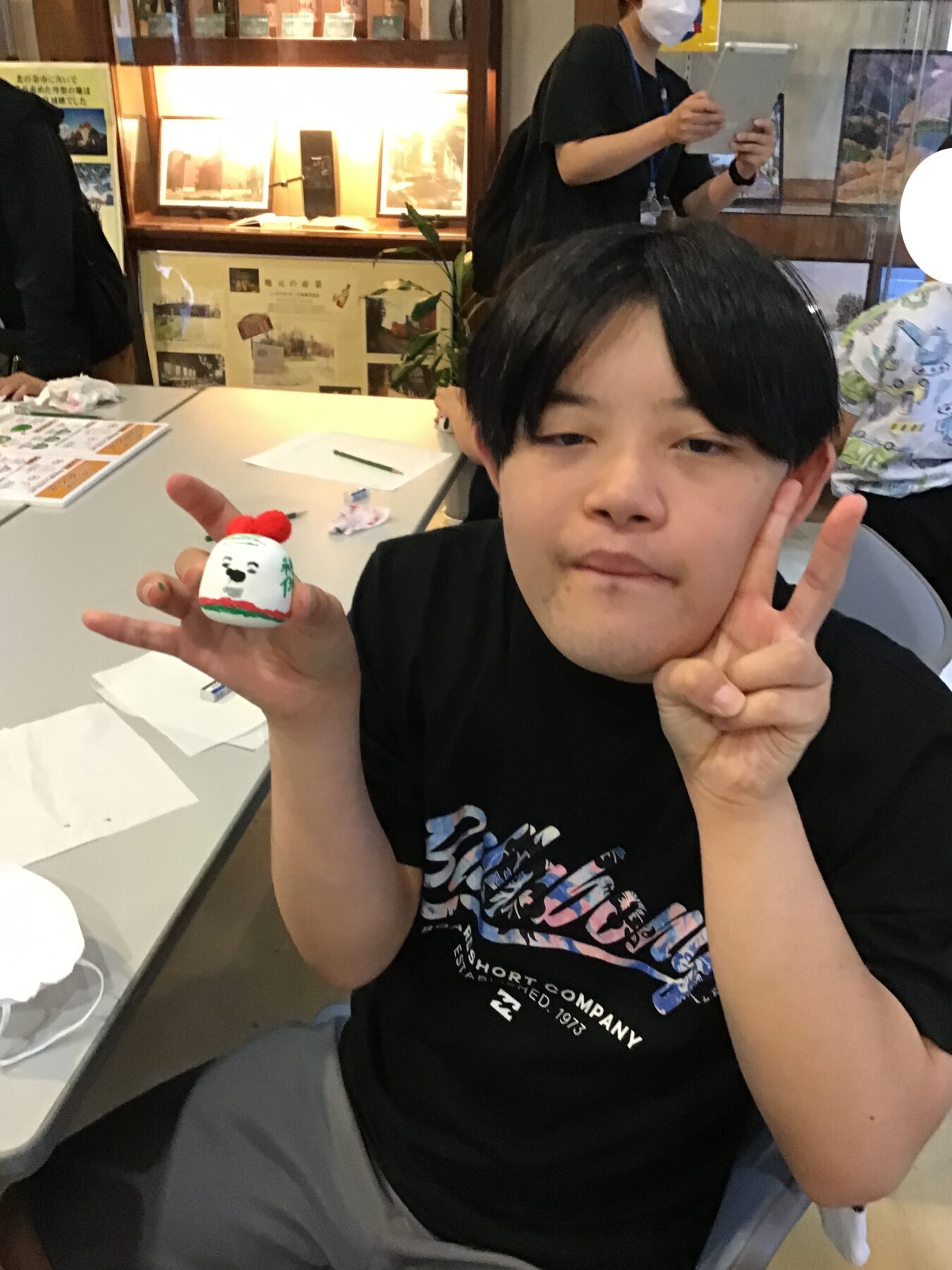 Young person with black hair wearing a black T-shirt sits at a table, holding a small white craft ball with a panda face and red pom-pom, making a peace sign.