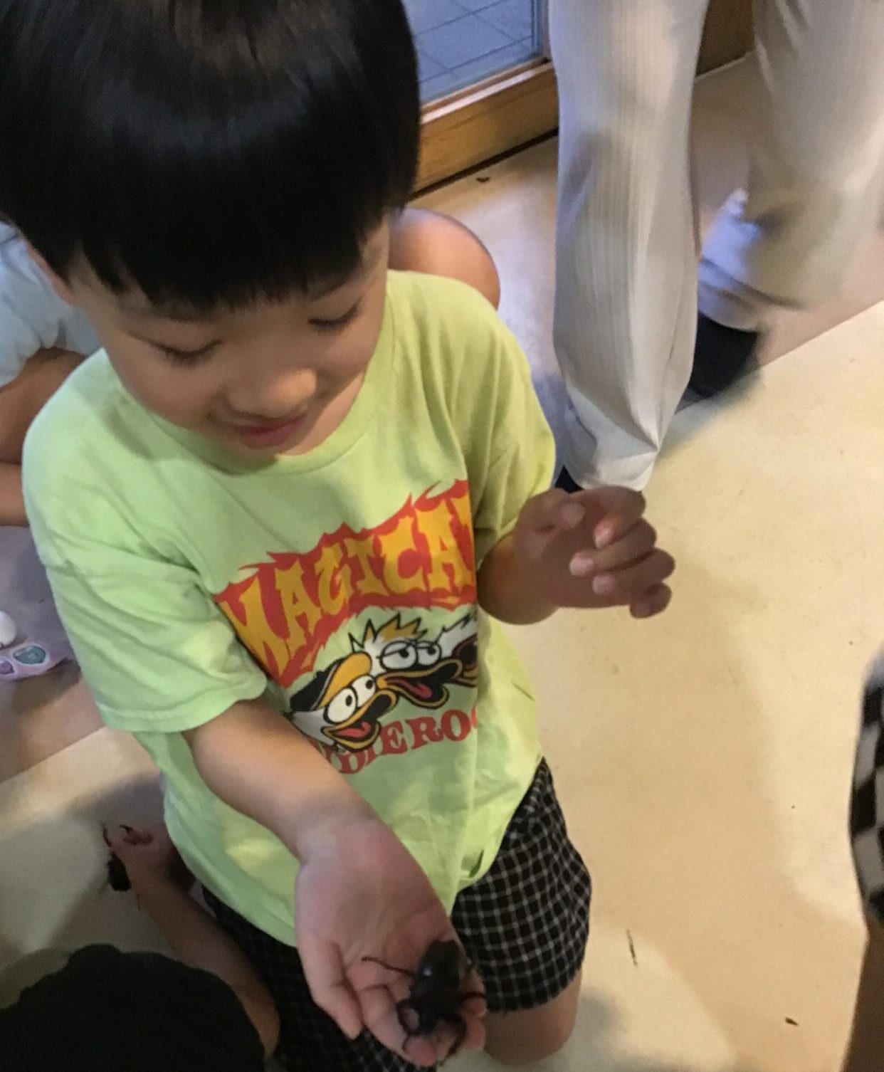 Young child in a lime T-shirt holds a small dark object in cupped hands, with others nearby indoors.