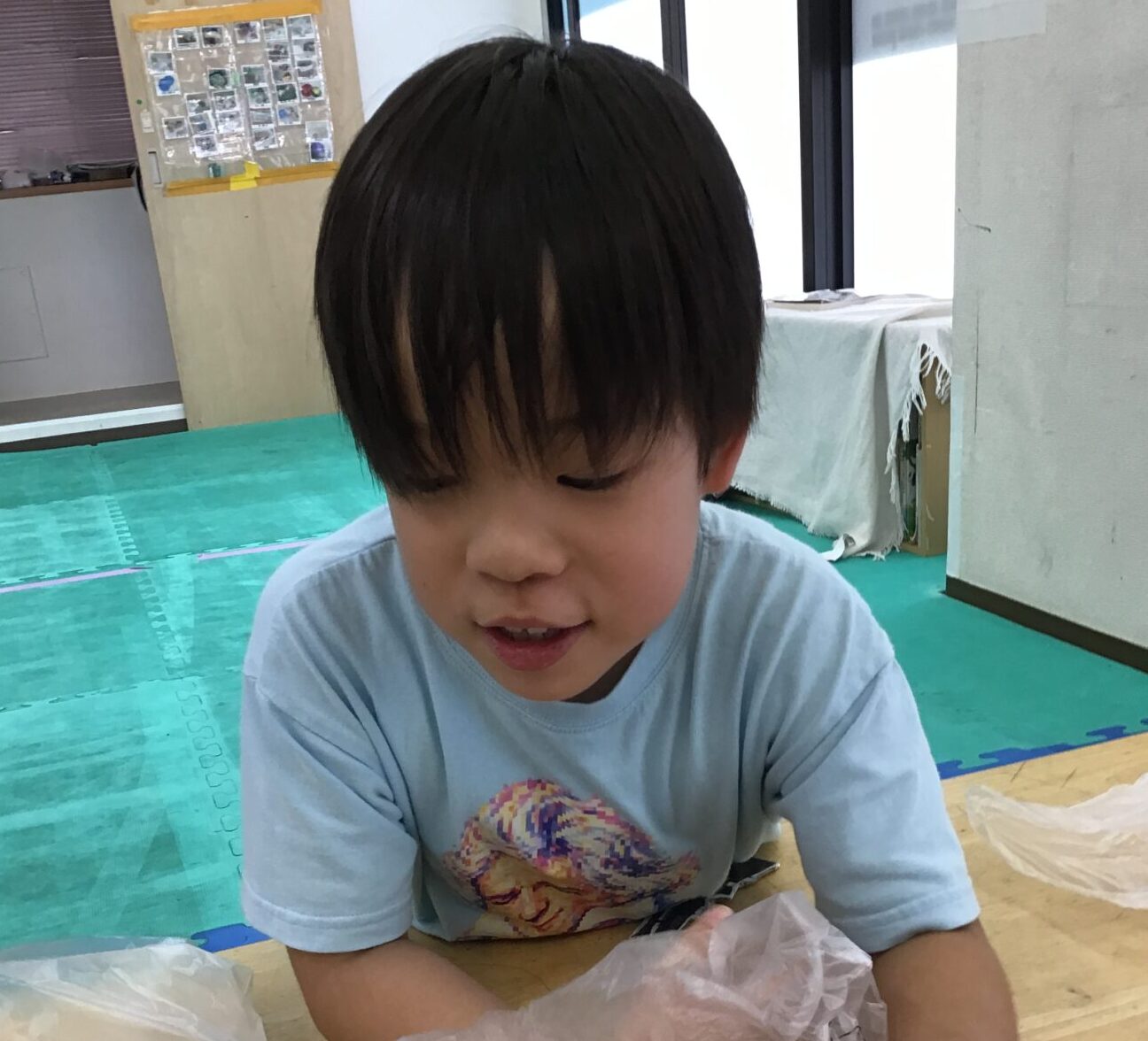 Young boy with dark hair sits at a table, handling a sheet of clear plastic wrap during a craft activity.