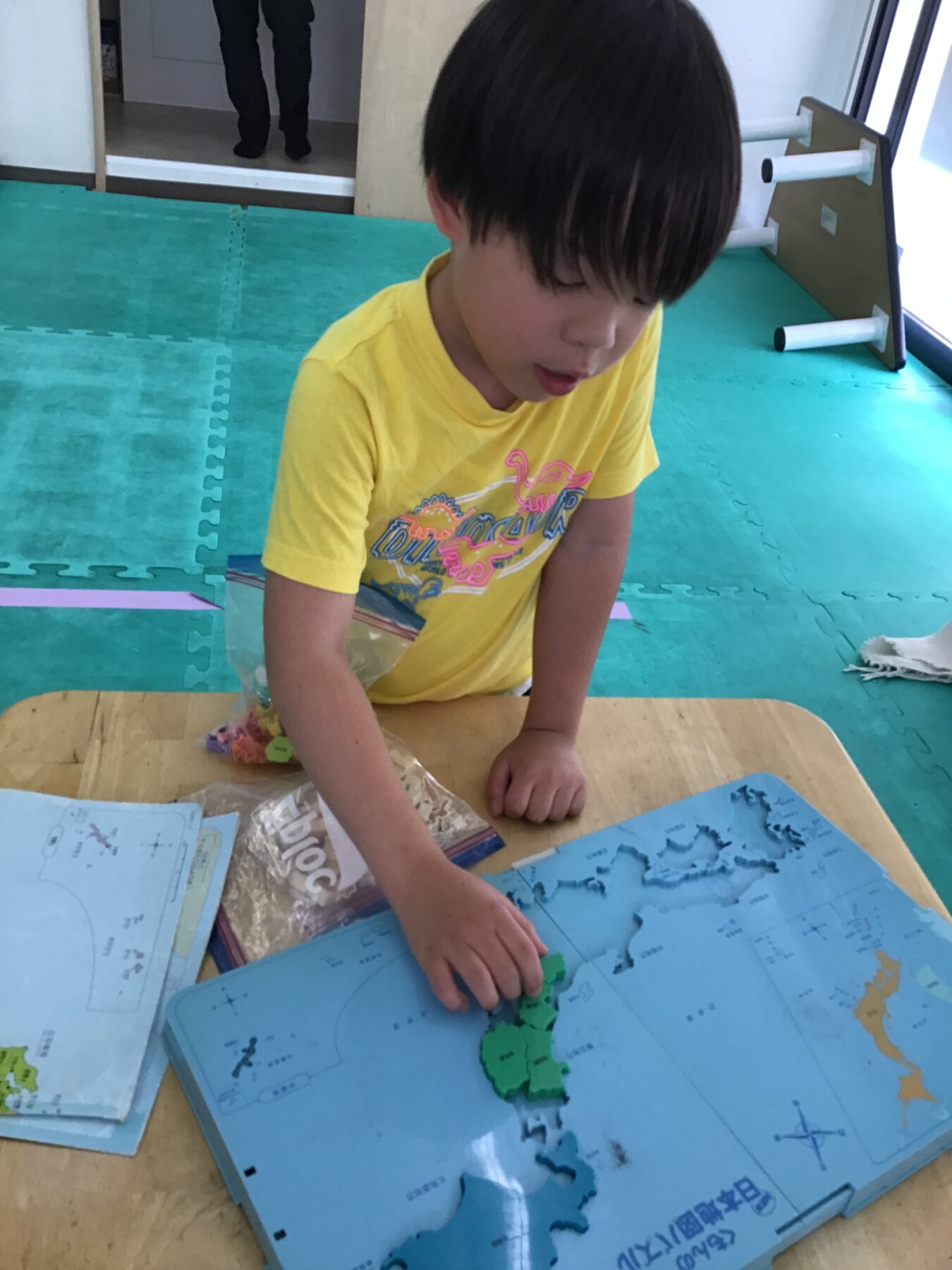 Young child in a yellow shirt works on a blue world map puzzle, placing green pieces on the table.