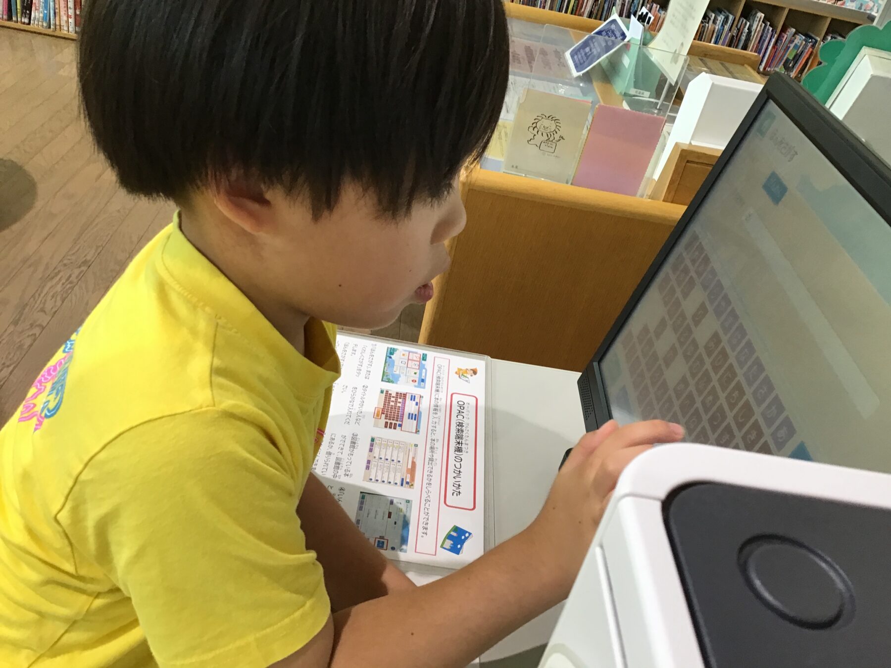 Young child in a yellow shirt using a touchscreen computer at a library desk with bookshelves in the background.