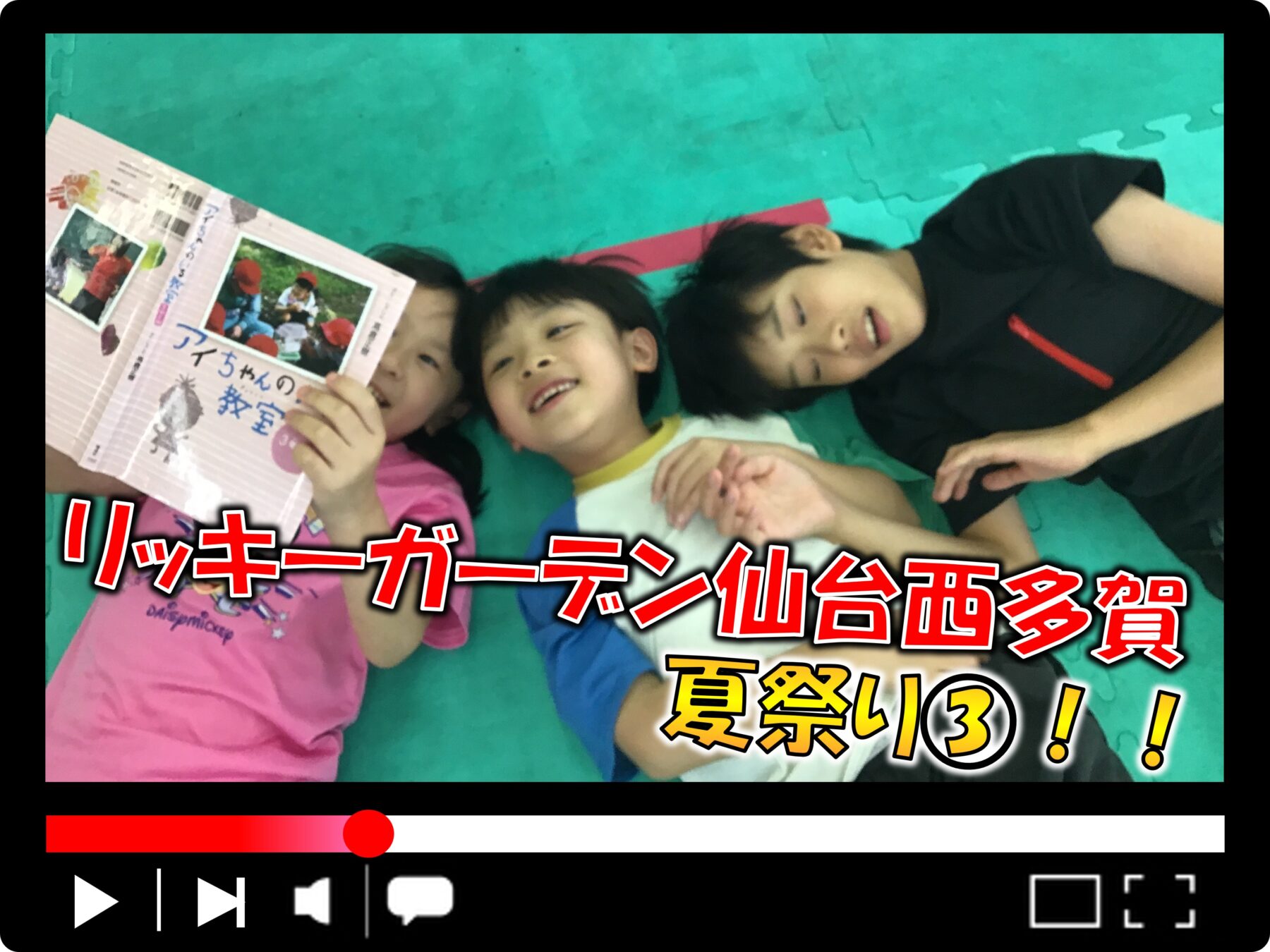 Three children lying on a green mat, one holding a pink photo book with Japanese text and smiling at the camera.