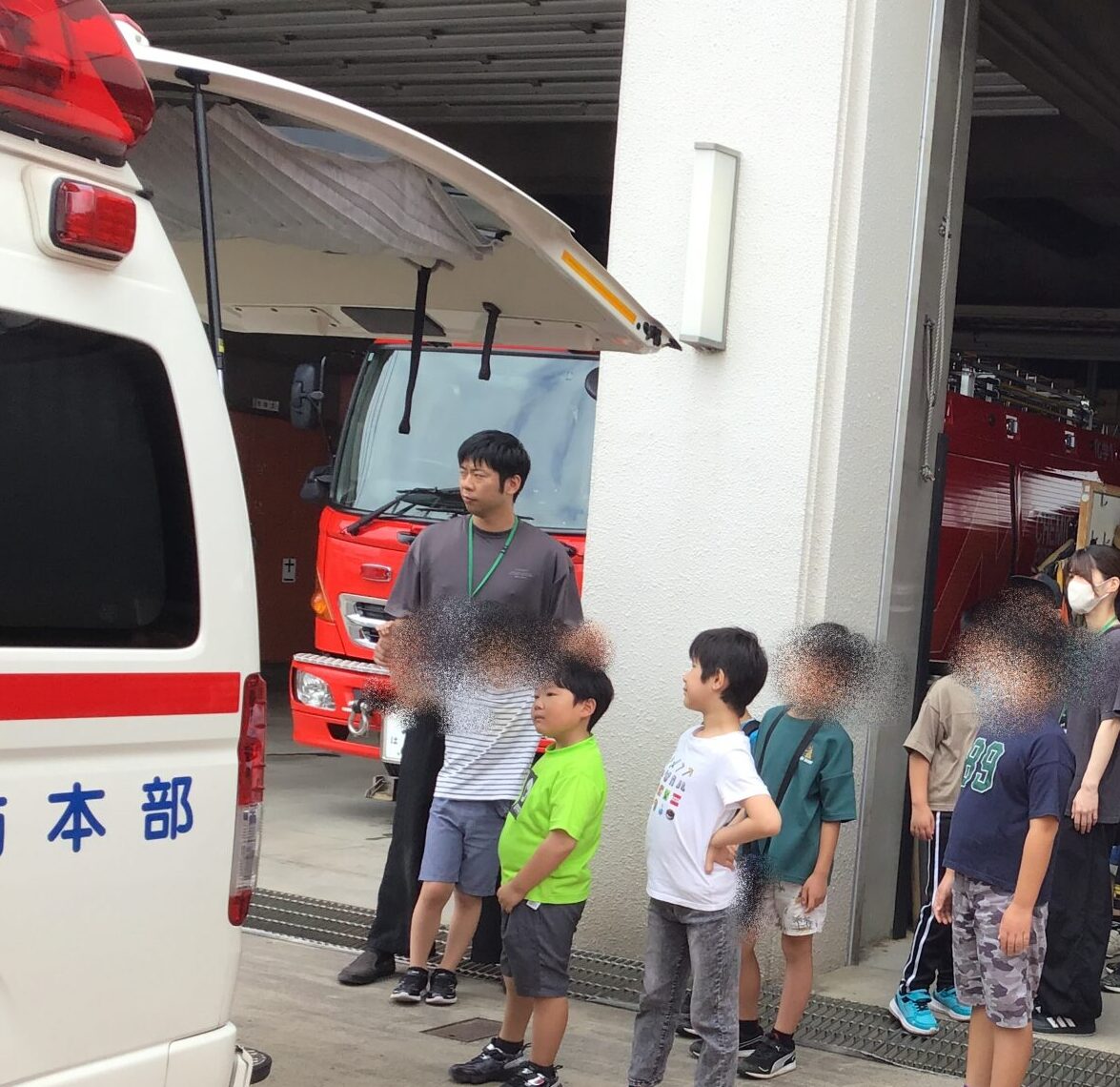 Line of children with backpacks and masks standing outside a fire station bay as red fire trucks sit inside behind them.