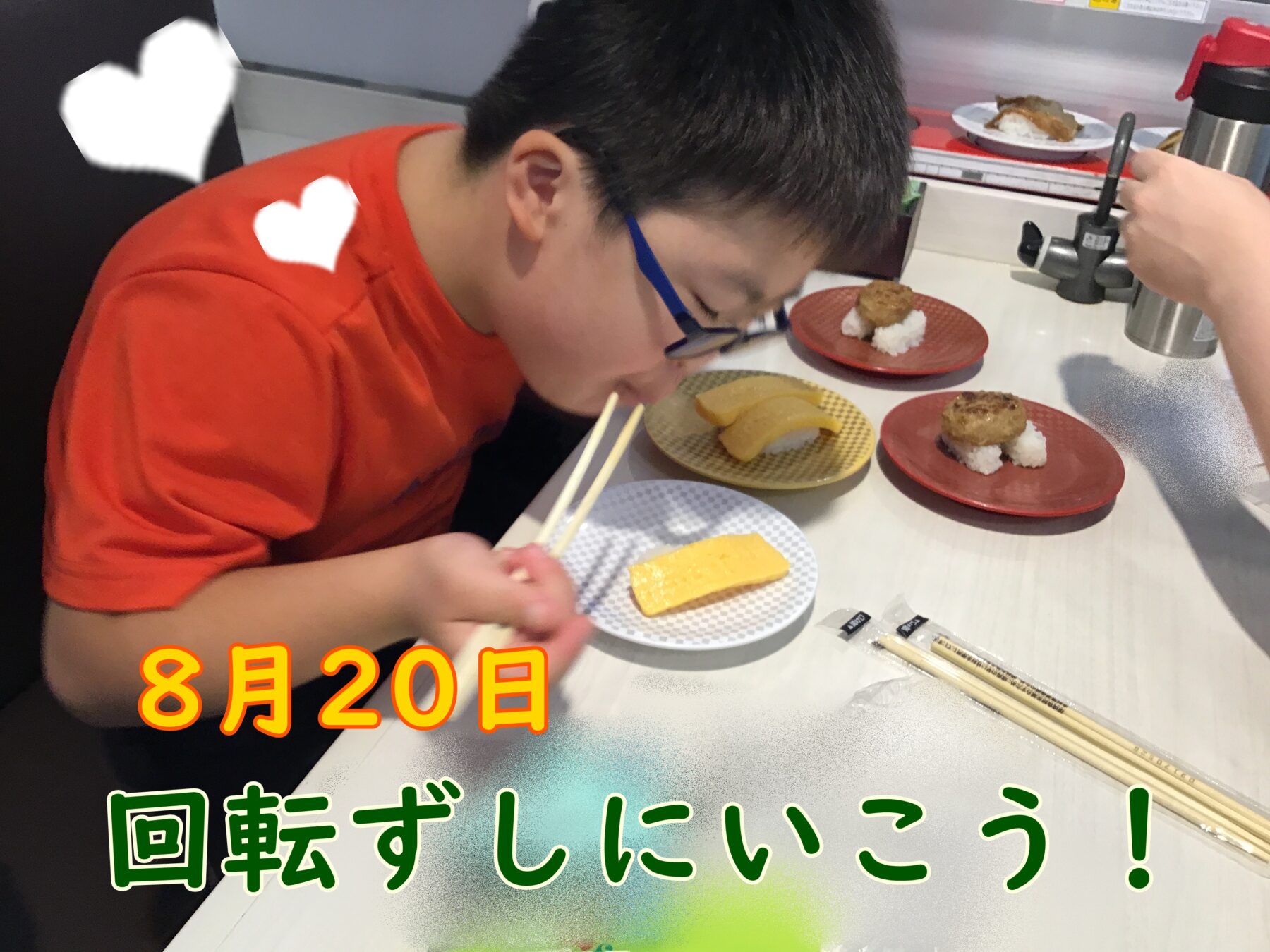 Young boy in an orange shirt eating with chopsticks at a sushi counter, plates of nigiri and tamagoyaki on the table, with a server's hand operating a metal lever in the background.