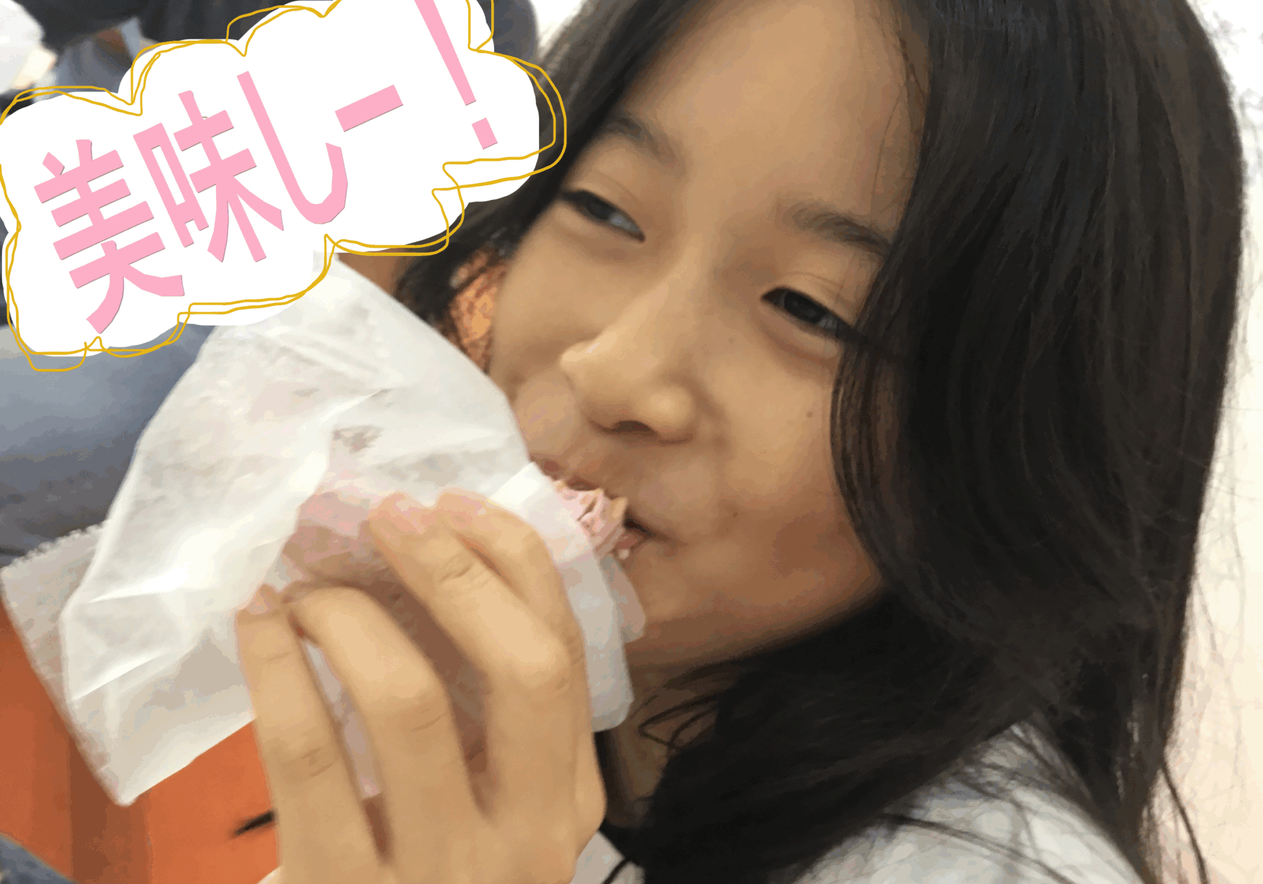 Girl smiling while biting into a pastry wrapped in white paper; a speech bubble with pink Japanese text 美味し! is drawn over the image.