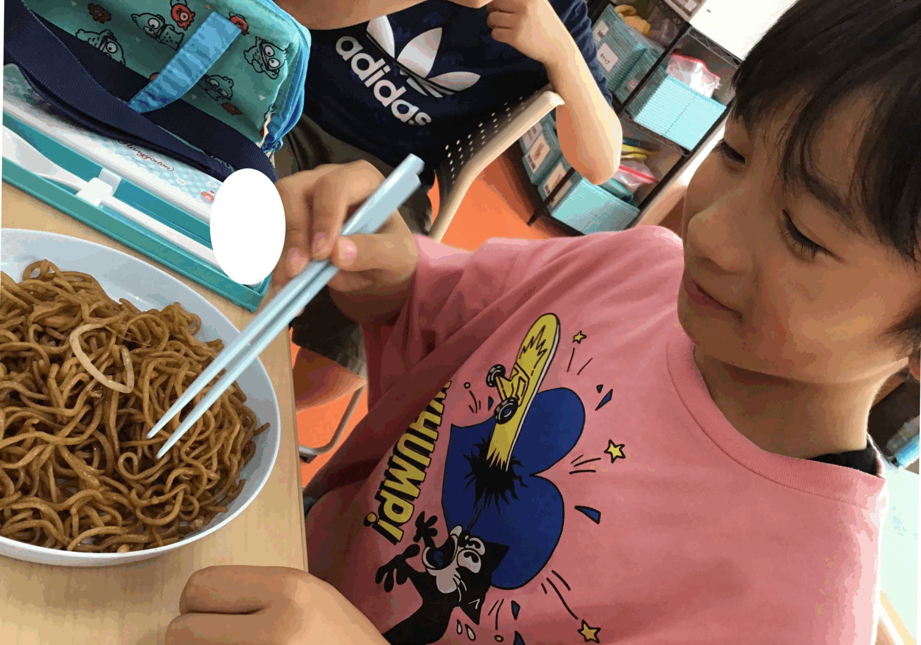 Child in a pink shirt uses blue chopsticks to pick up noodles from a bowl at a cafeteria table