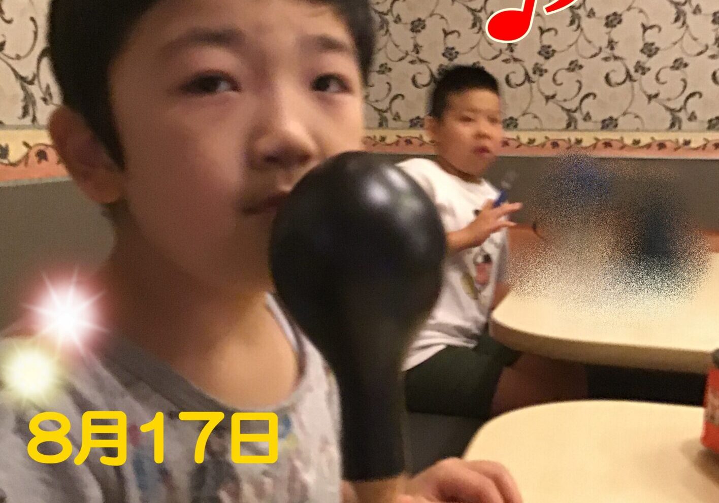 Close-up of a person in the foreground in a karaoke venue, with another person seated at a table in the background and a large red musical note overlay plus a date stamp.