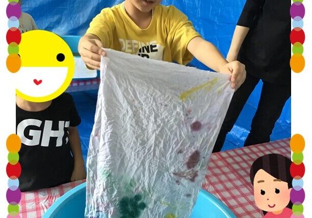 Young boy in a yellow shirt holds up a wrinkled white cloth smeared with green, purple, and yellow paint over a blue tub at a kids' art station.