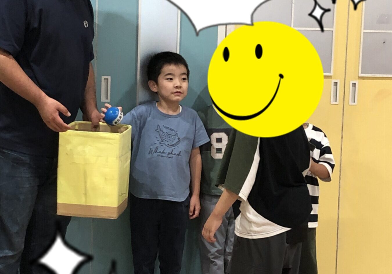 Man wearing a mask hands a yellow box to a standing boy as others watch in a colorful classroom setting.