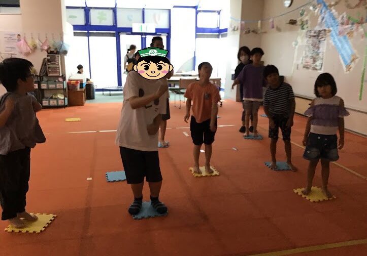 Group of children standing on colored foam tiles arranged in rows inside a bright classroom, ready for a game or activity. One child in the foreground has a cartoon sticker over their face for privacy.