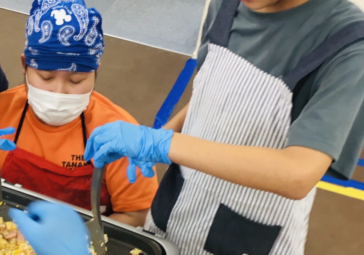 Two kids wearing blue bandanas and aprons cook fried rice in a portable pan on a table, with one stirring the dish.