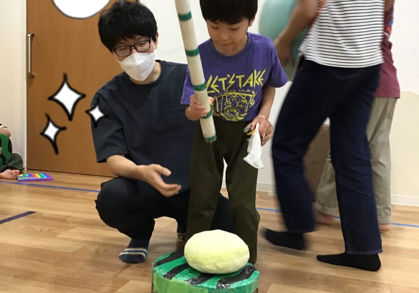 Child in a purple shirt holds a long striped baton, aiming at a foam target atop a taped green-and-brown stacked cylinder, while an adult in a mask crouches nearby; speech bubble reads よーし！
