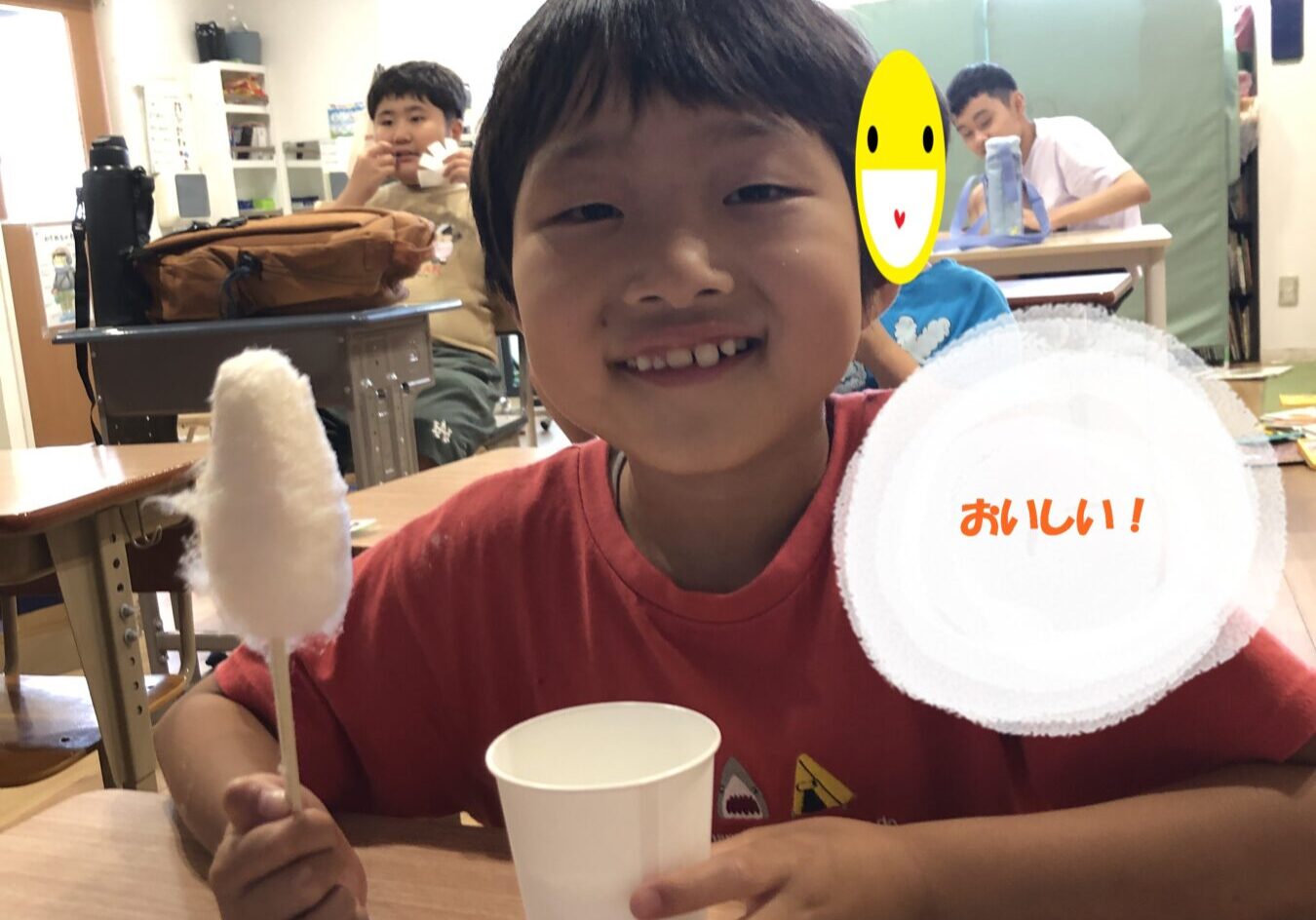 Smiling boy in a red shirt at a classroom table holding cotton candy on a stick, with a white cup; background kids and a circular overlay reading 'おいしい！'