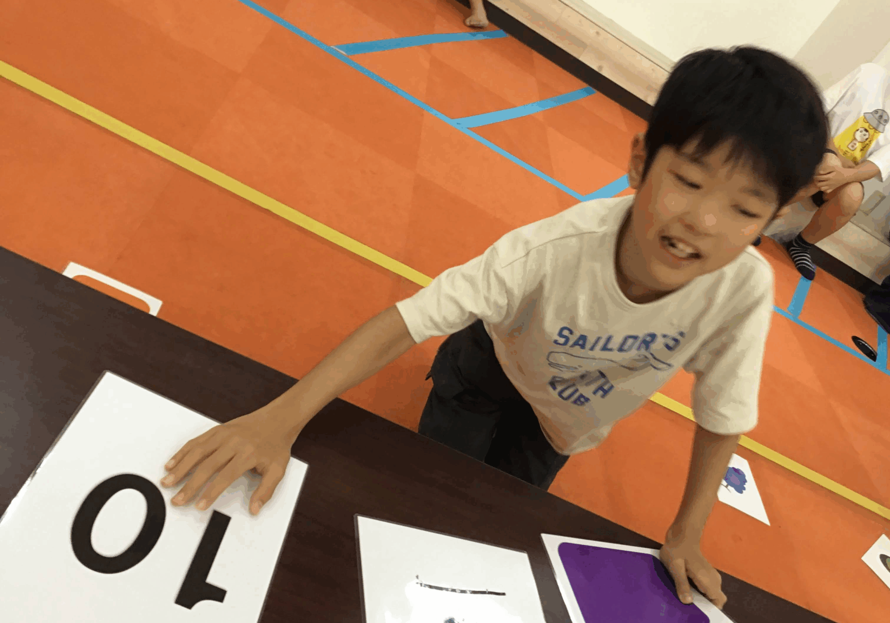 A boy leaning over a table places a large white card with bold black numbers during a classroom activity on an orange gym floor with blue tape lines in the background.