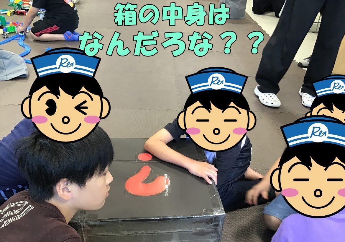 Children sit and kneel on a foam mat around a taped cardboard box, a red curved toy on top, with Japanese text asking what's inside.