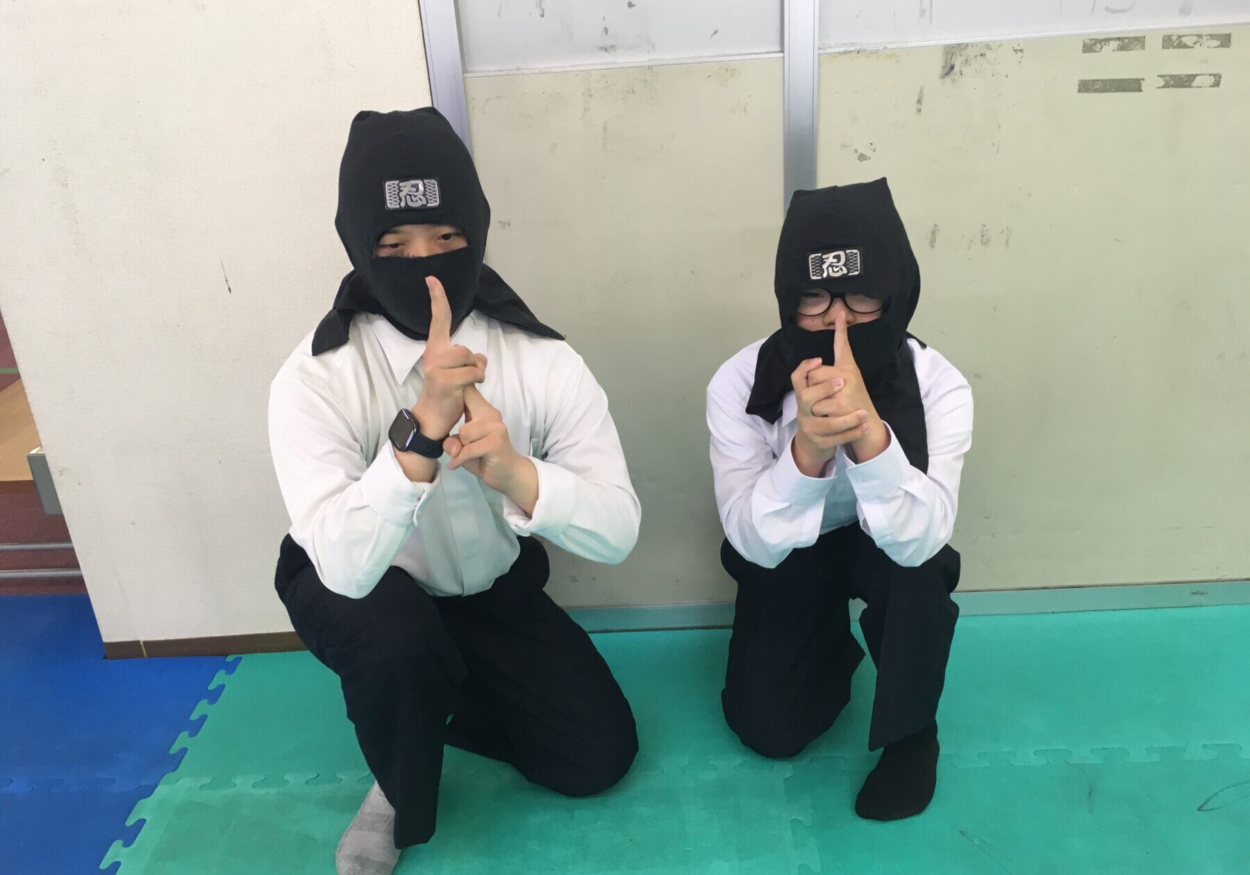Two people in ninja costumes kneel on a colorful foam mat, making a shushing gesture with a finger to the lips (patch that reads 忍 on their hoods).