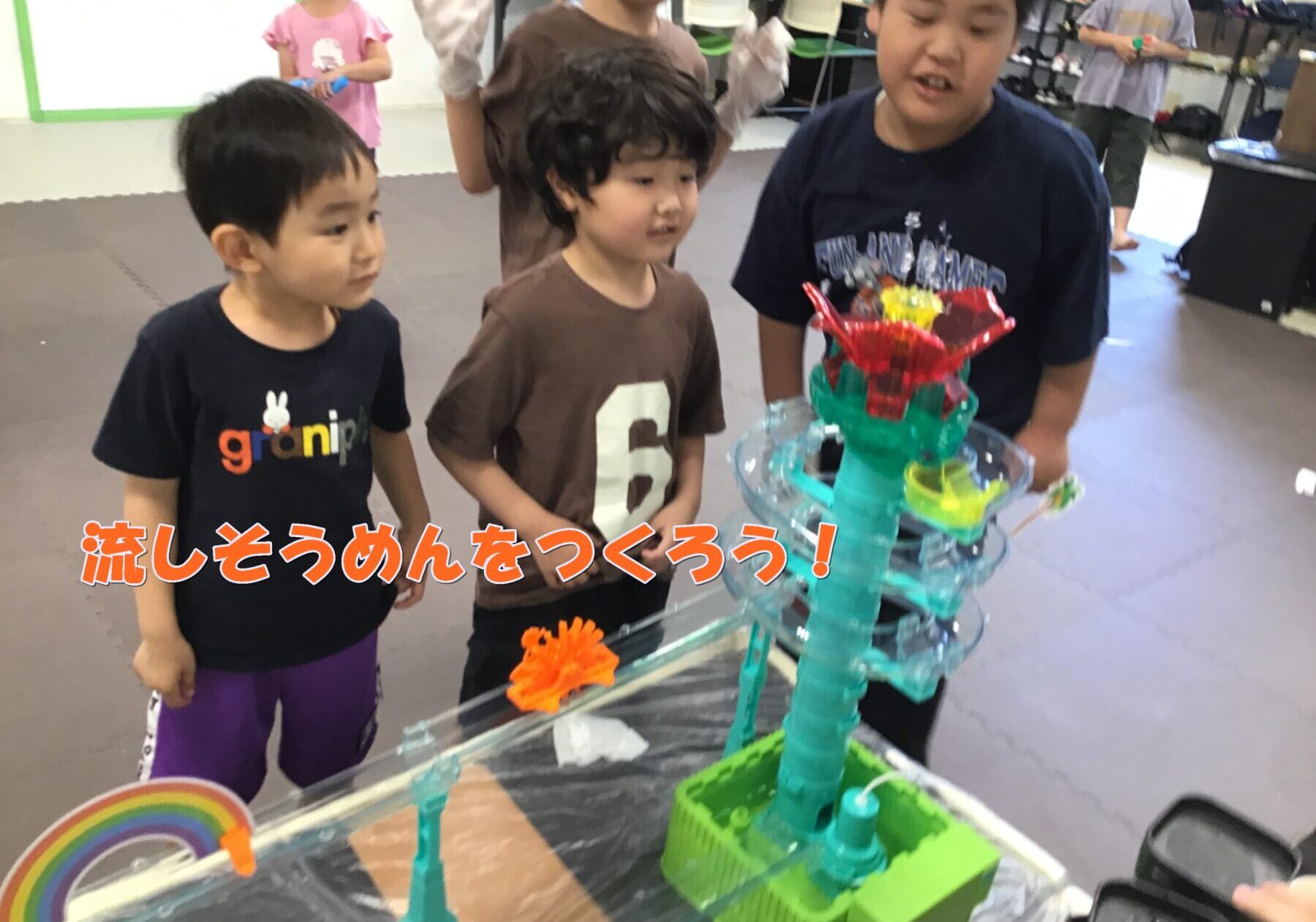 Three children watch a colorful DIY water-flow sculpture during a nagashi somen activity in a classroom setting